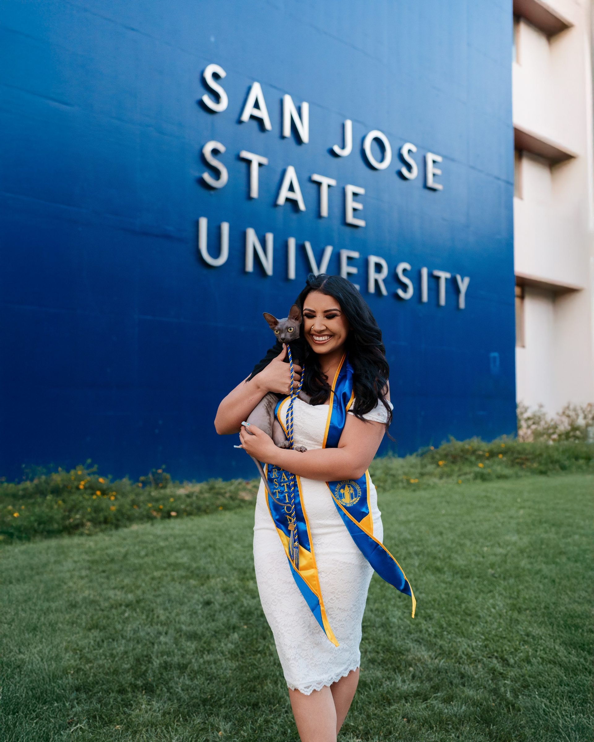 Maya laughing and cuddling her sphynx cat wrapped in a pink blanket in front of the SJSU campus sign