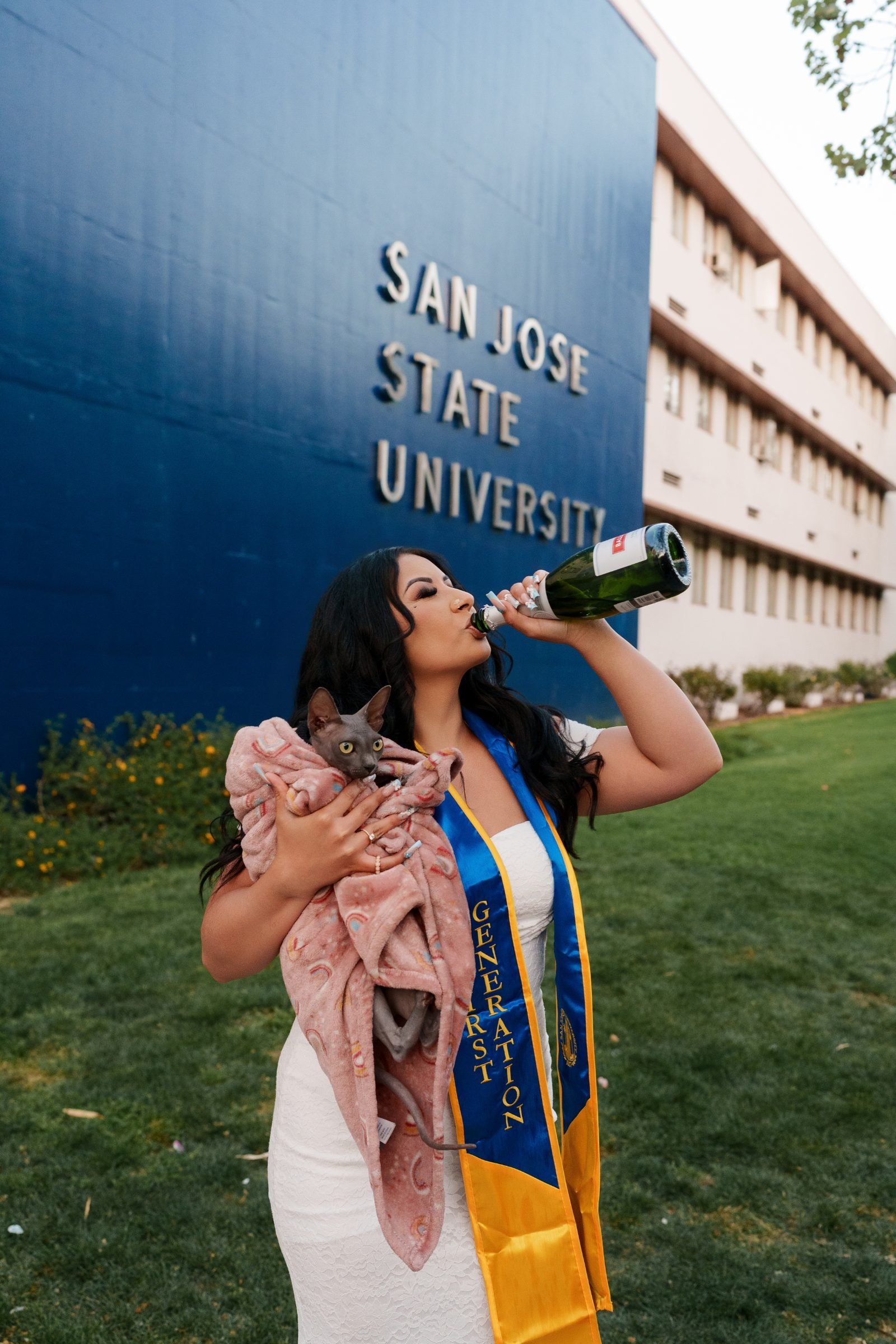 Maya celebrating in her First Generation sash and white dress, champagne bottle raised at the San Jose State University sign