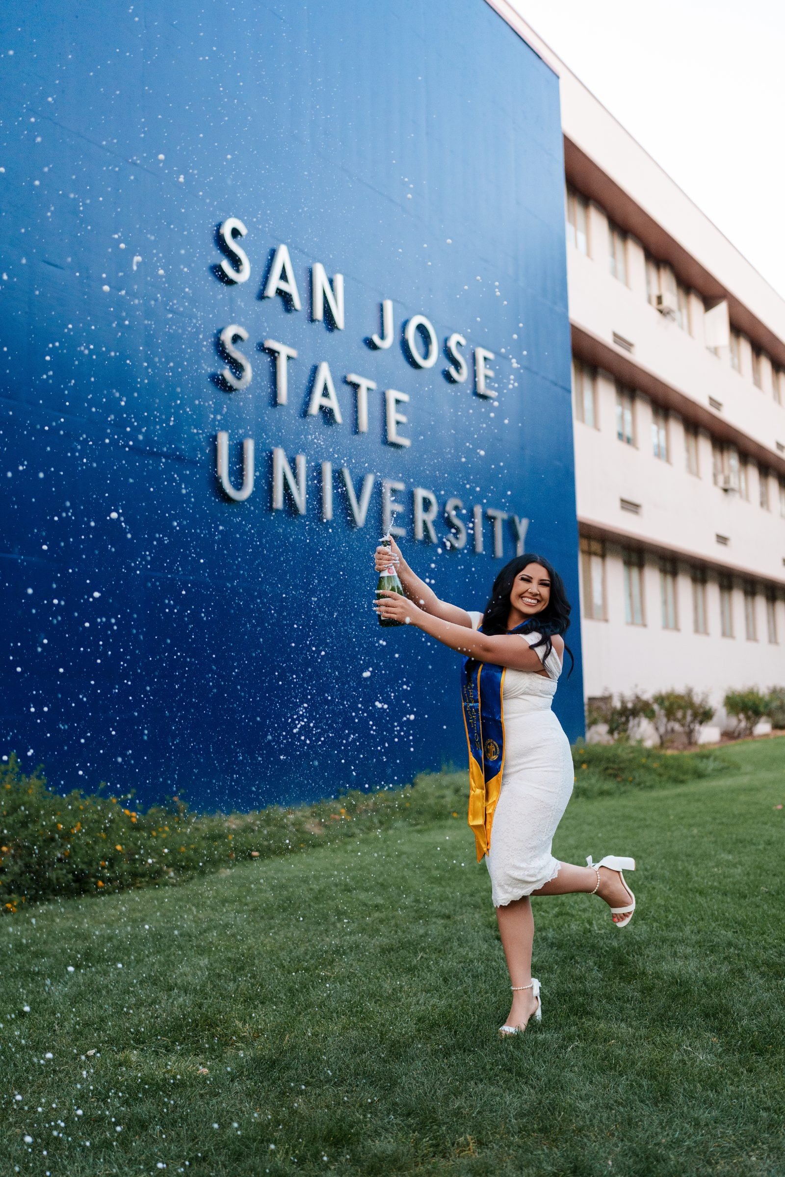 Maya popping a champagne bottle with mist spraying across the blue San Jose State University campus wall