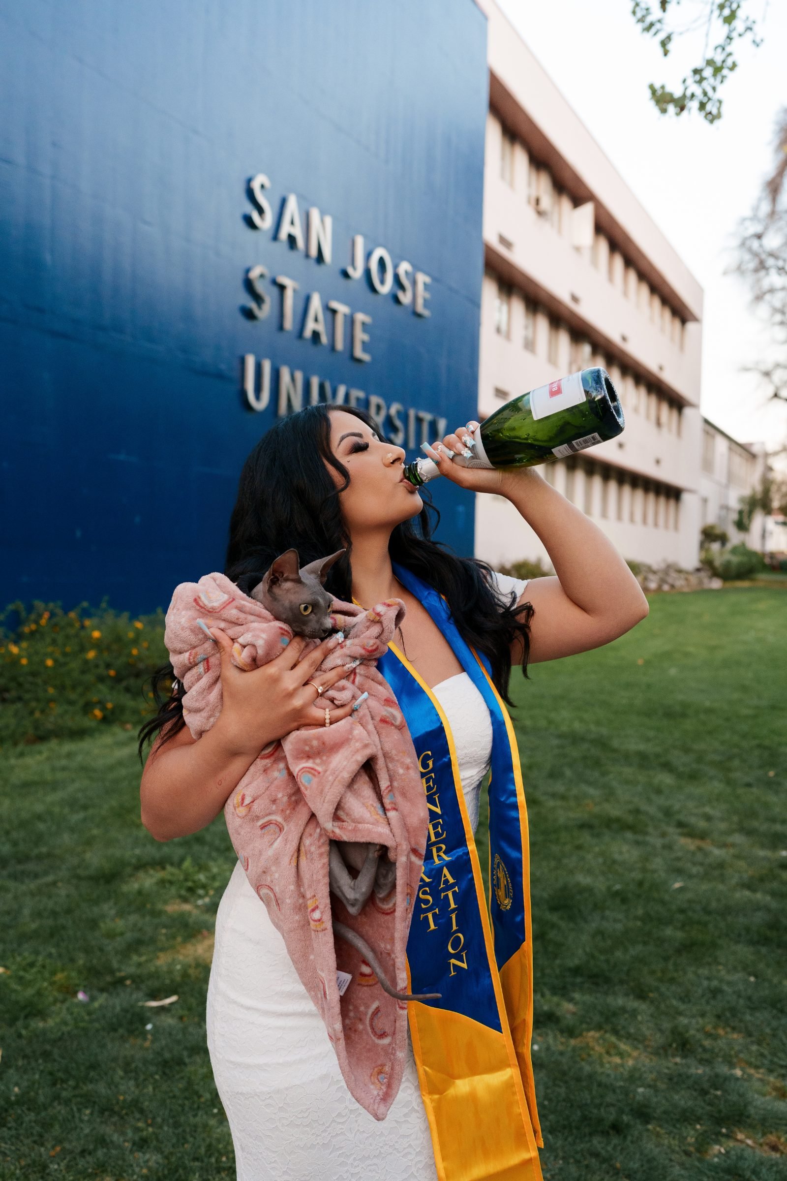 Maya holding her blanketed sphynx cat and drinking from a champagne bottle in front of the SJSU sign