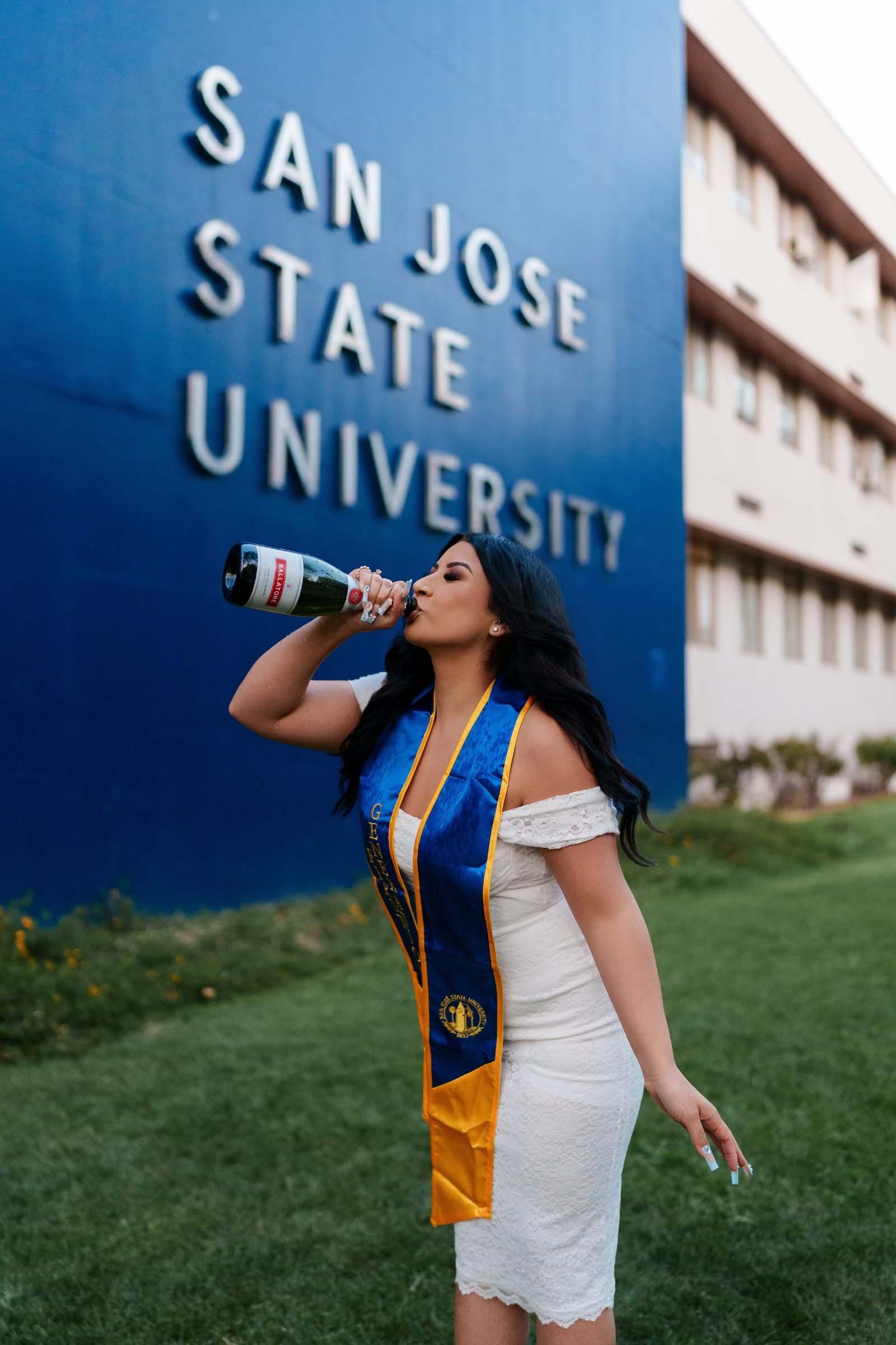 Close-up of Maya in white lace dress and SJSU sash drinking champagne alone in front of the San Jose State University sign