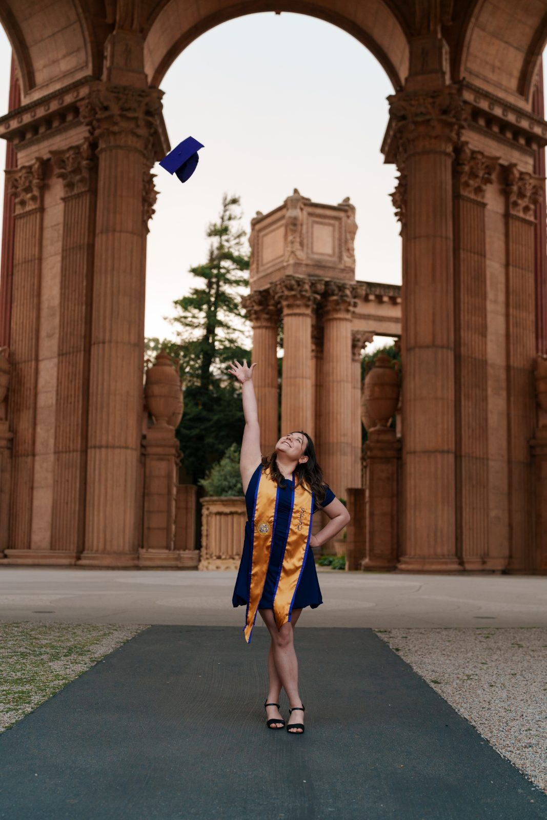 SFSU graduate in navy dress and gold sash tossing purple cap under the Palace of Fine Arts colonnades, San Francisco
