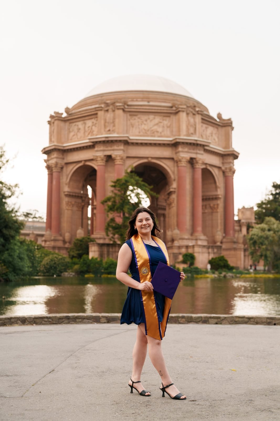 SFSU grad holding purple mortarboard cap in front of the Palace of Fine Arts rotunda and lagoon, San Francisco