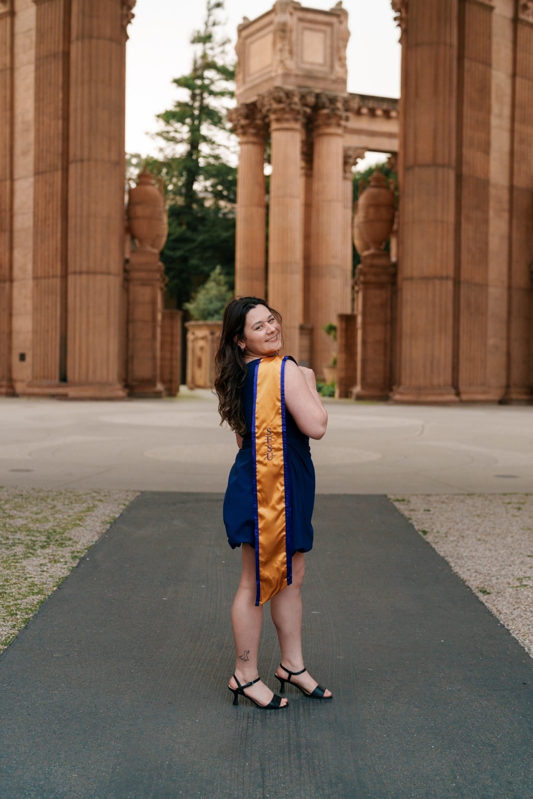 Graduate in SFSU gold sash looking over her shoulder between the sandstone columns of the Palace of Fine Arts colonnade