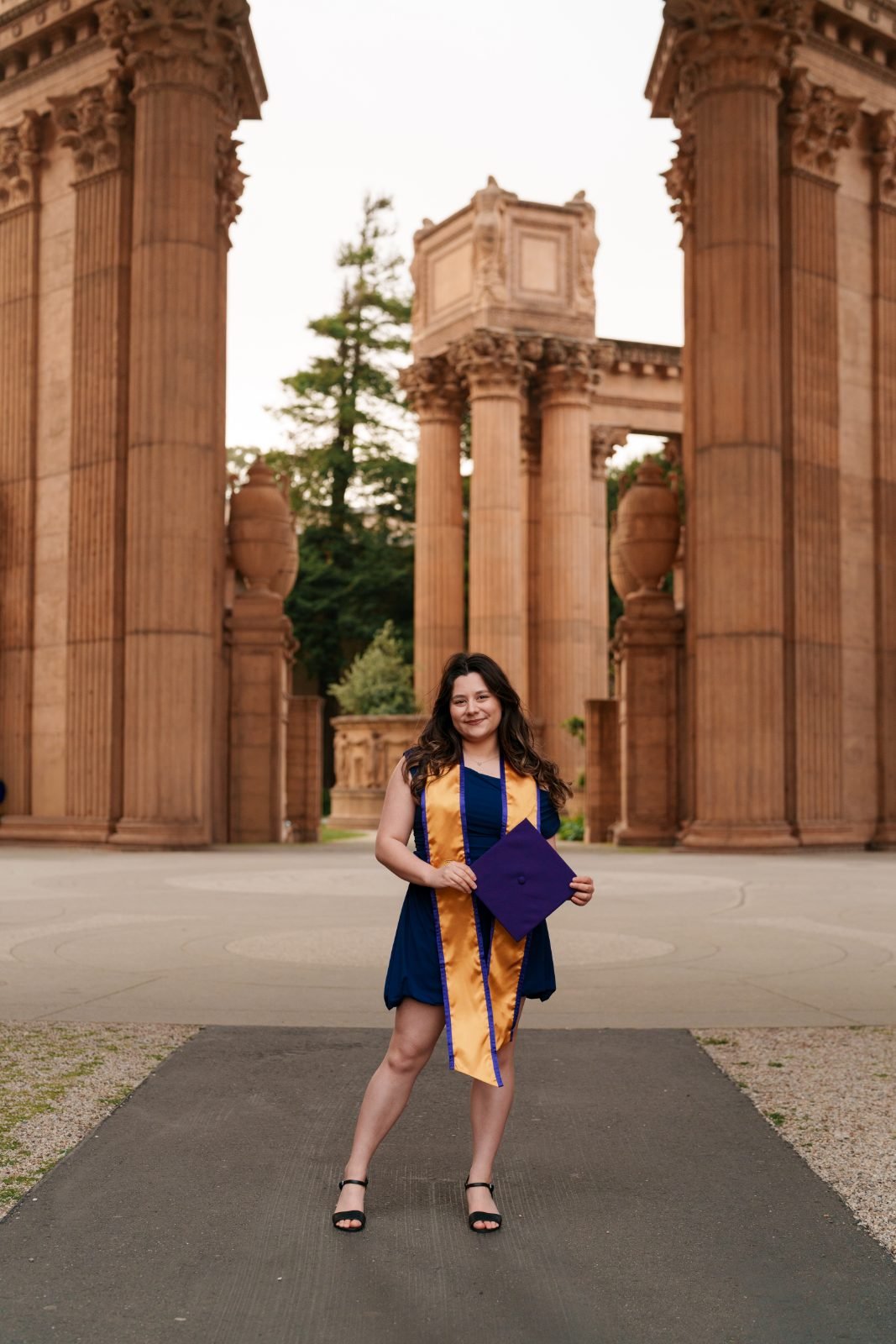 SFSU graduate in navy dress holding purple cap and smiling between towering Palace of Fine Arts columns