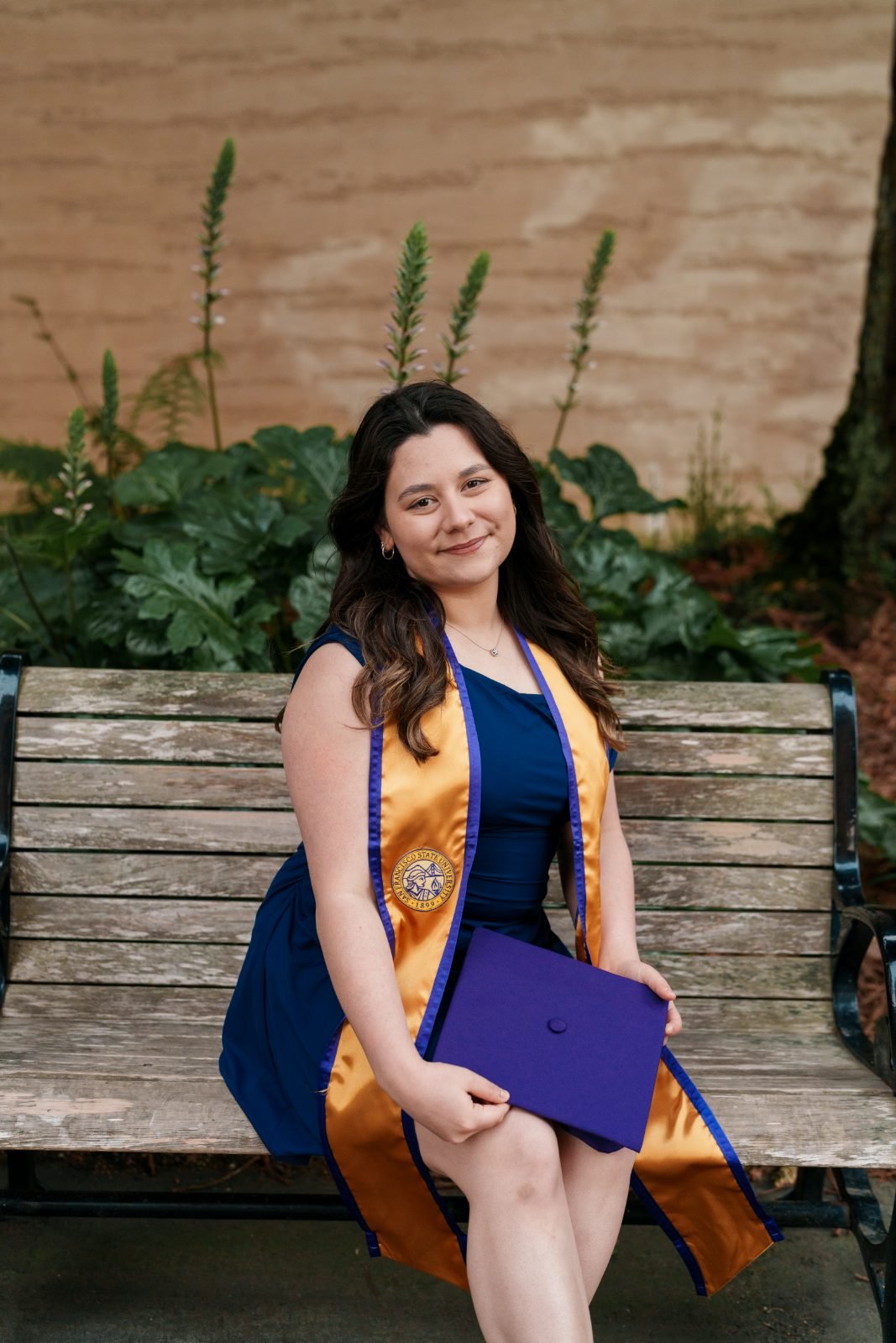 SFSU grad sitting on a wooden bench holding her purple mortarboard, soft garden greenery in the background