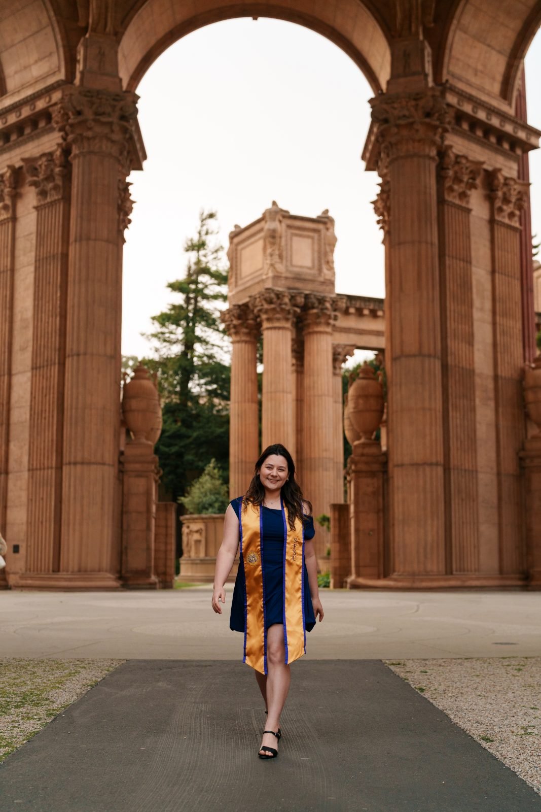 Graduate in SFSU gold and navy sash walking confidently through the Palace of Fine Arts arch toward the camera