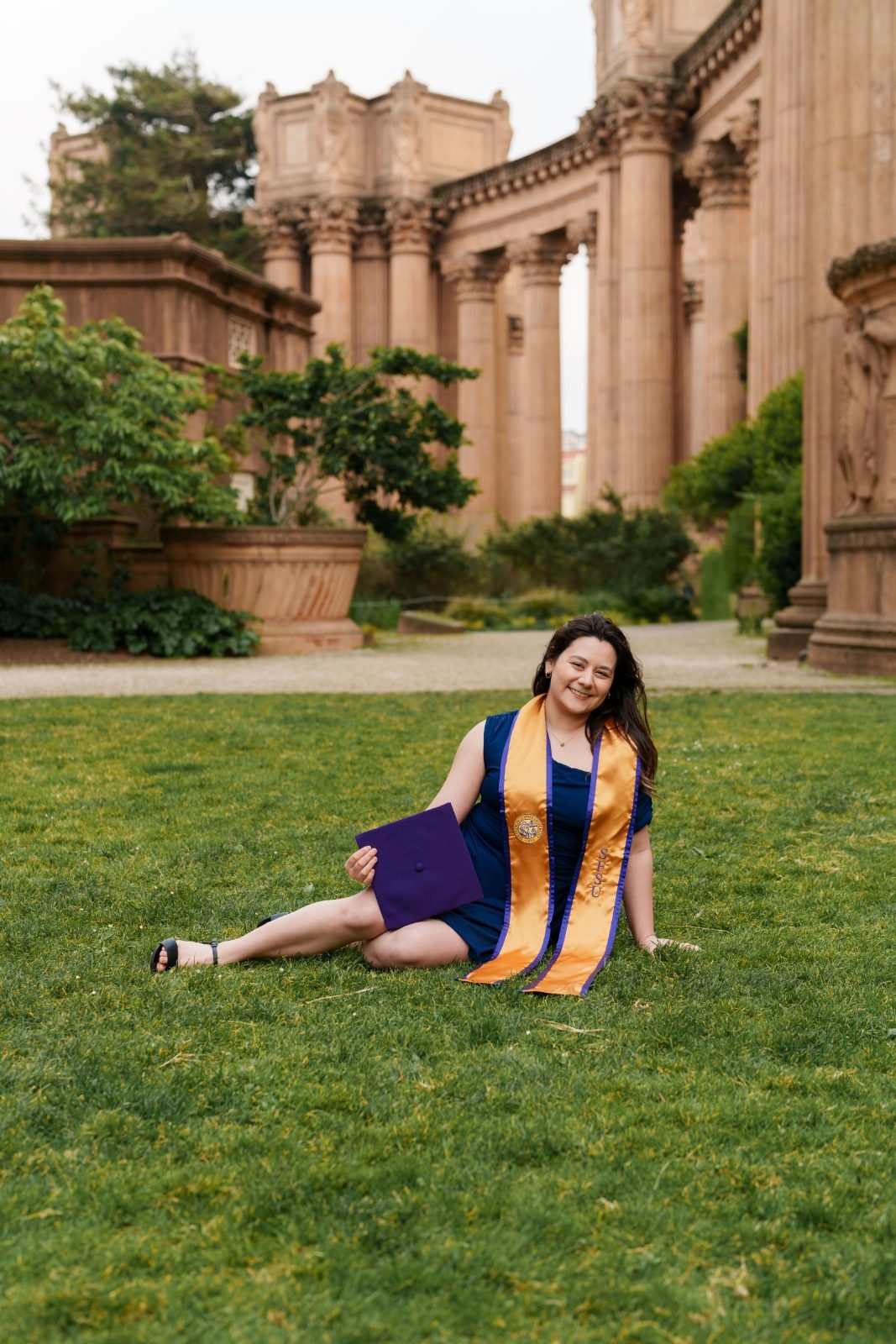 SFSU grad sitting on the grass beside the Palace of Fine Arts colonnade, smiling with purple cap resting in her lap
