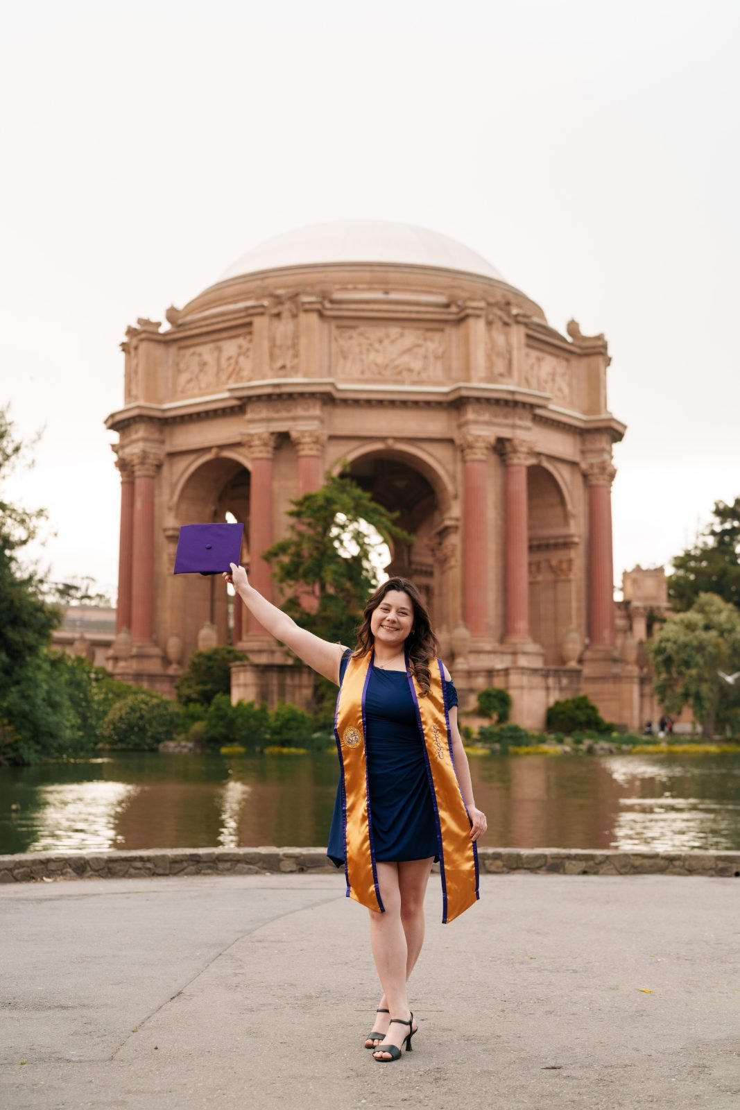SFSU graduate raising her purple mortarboard in front of the Palace of Fine Arts rotunda dome and lagoon