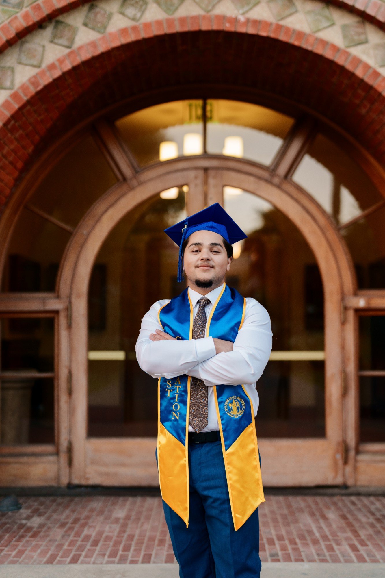 SJSU grad in cap and blue and gold stole stands arms crossed under the Romanesque arched brick entrance