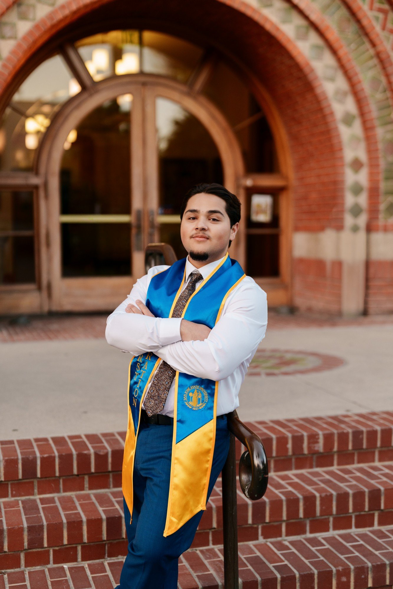 SJSU graduate with arms crossed in blue and gold stole leans against a railing outside the arched brick campus building