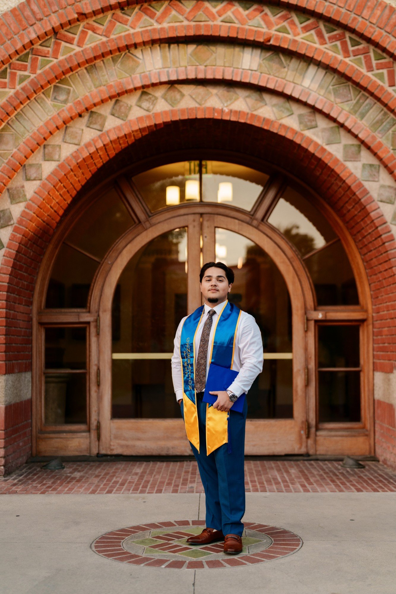 SJSU graduate holding his diploma folder in front of the ornate arched brick and tile entrance of the campus Student Union