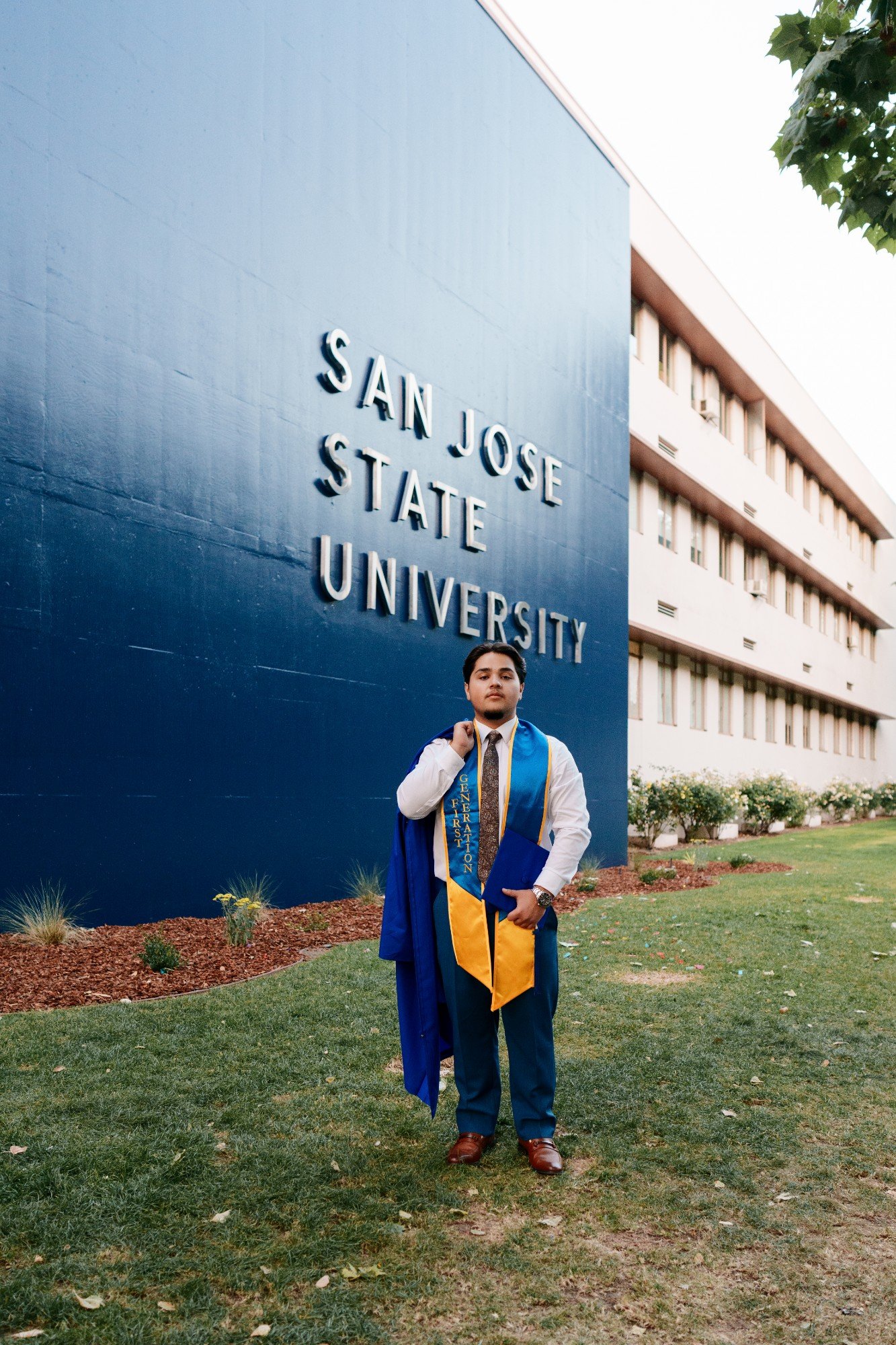 SJSU grad in navy suit, blue and gold stole, and graduation gown poses in front of the San Jose State University sign
