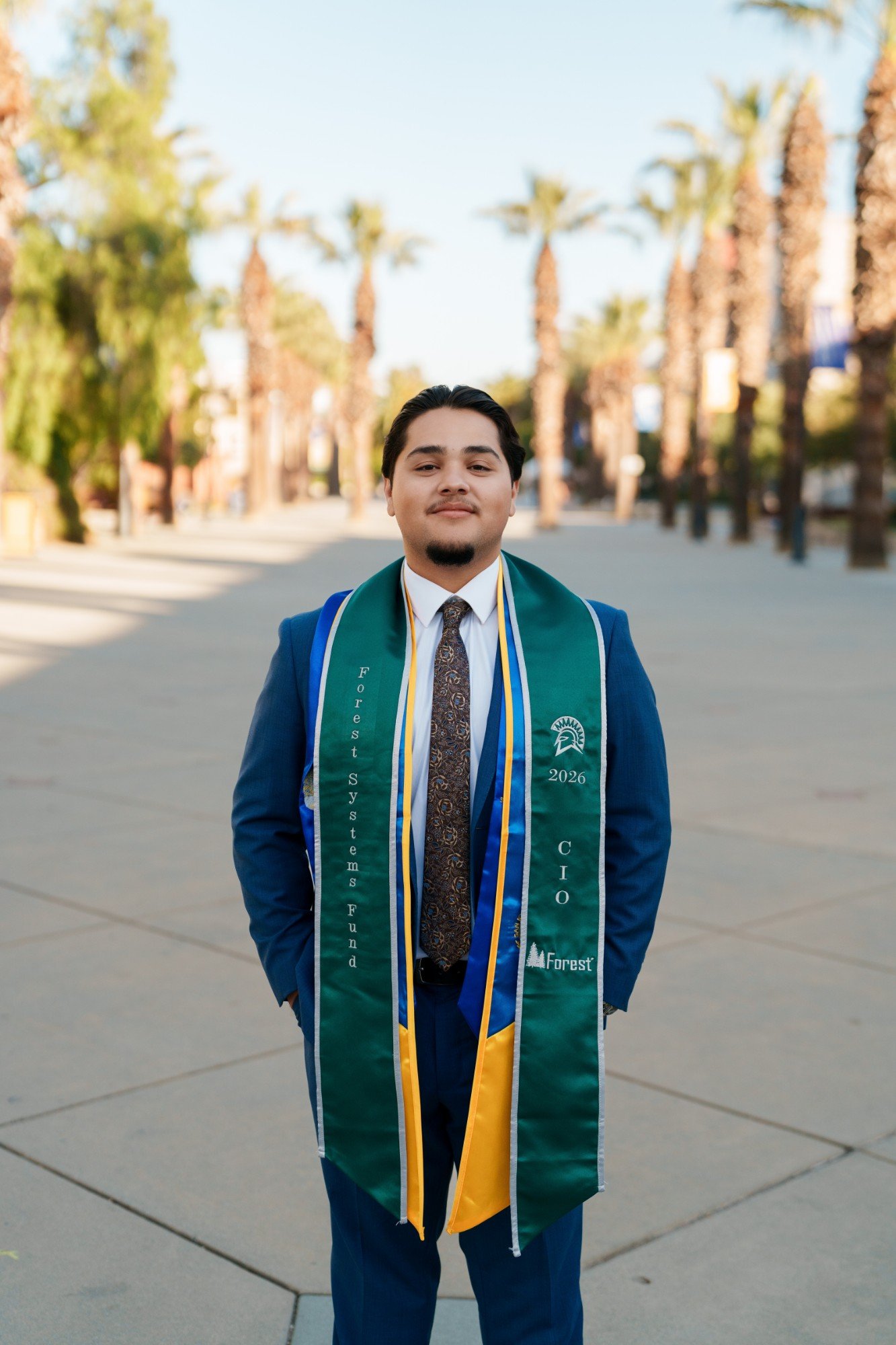 SJSU grad smiling on the palm tree promenade in a blue suit with green and gold graduation stoles layered