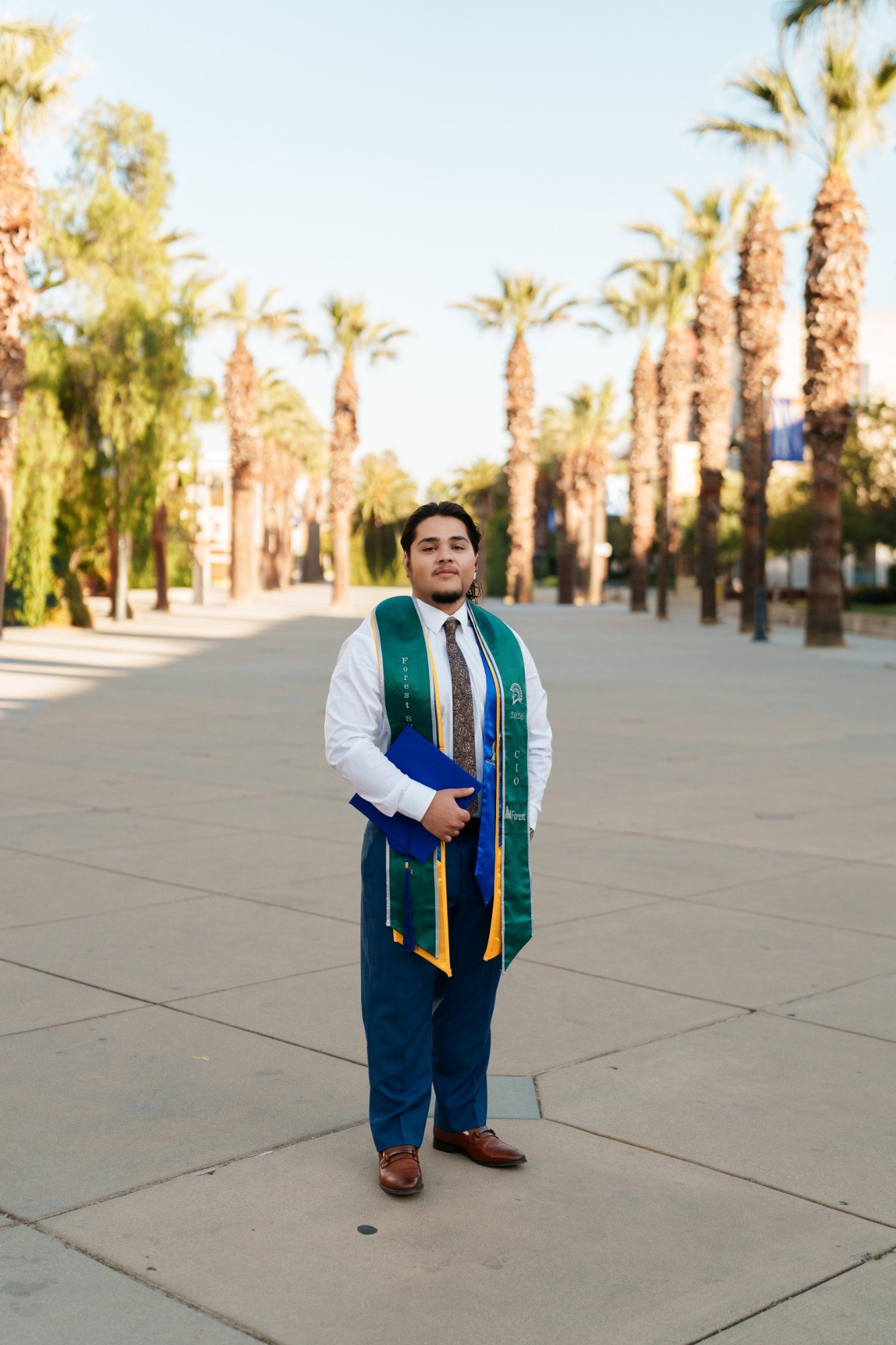 Full-length portrait of SJSU graduate holding his cap on the palm-lined campus promenade with green and gold stoles
