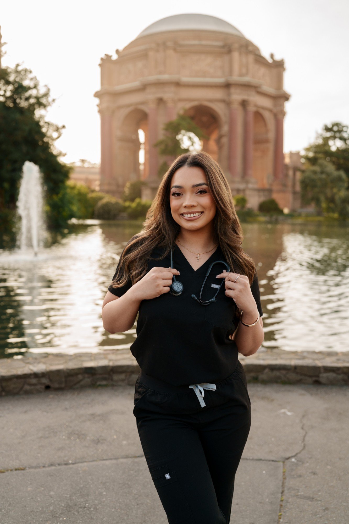 SMU nursing grad in black scrubs with stethoscope smiling in front of Palace of Fine Arts rotunda and lagoon fountain
