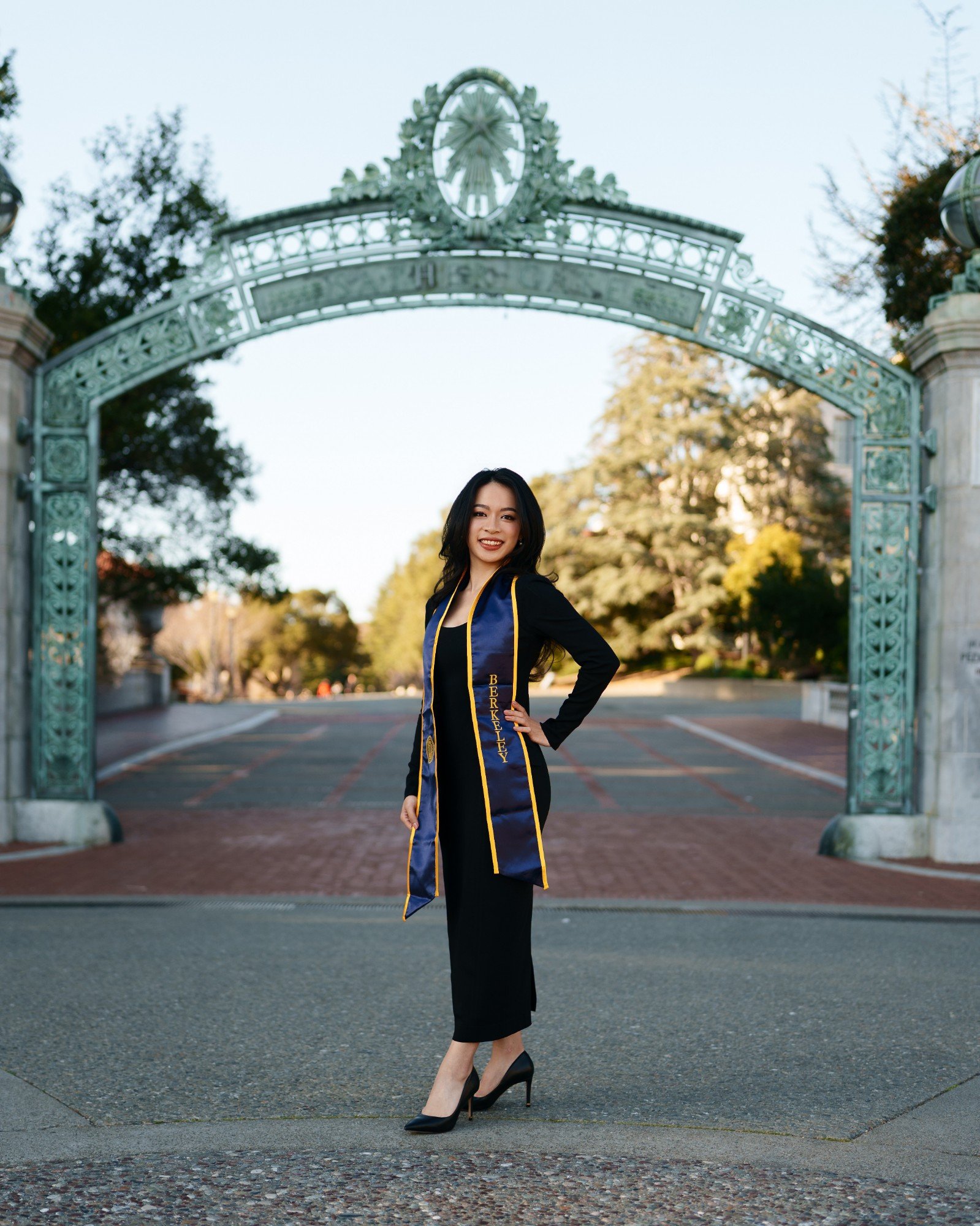 UC Berkeley graduate in black dress and navy-and-gold Berkeley stole standing centered beneath Sather Gate on a clear winter