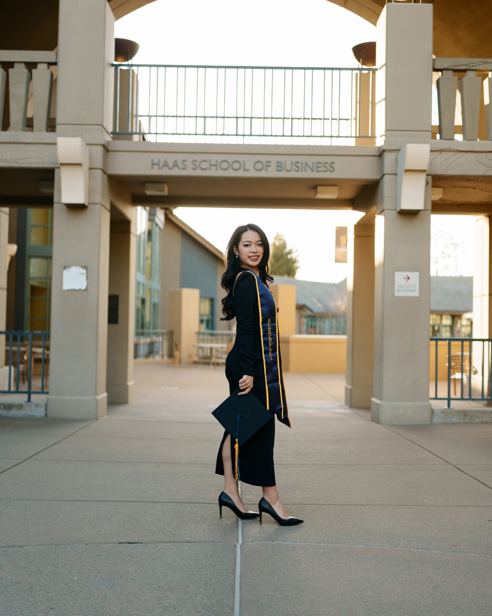 UC Berkeley graduate walking away from camera holding mortarboard at her side at the Haas School of Business courtyard entran