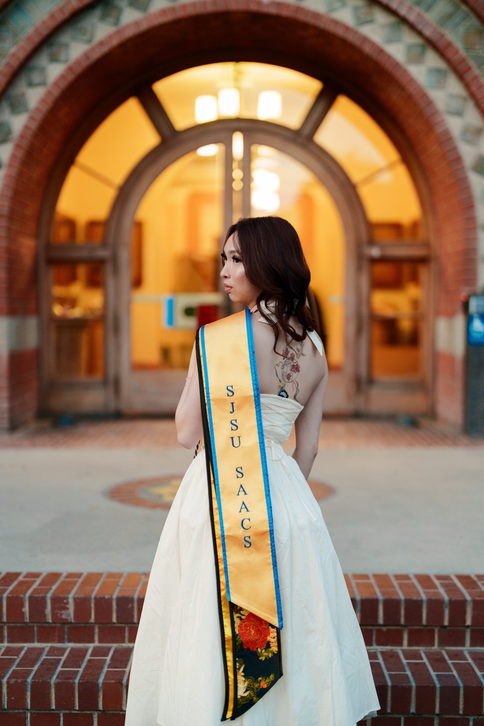 SJSU SAACS graduate in white backless dress and floral-embroidered sash posing in front of the glowing arched doorway of the