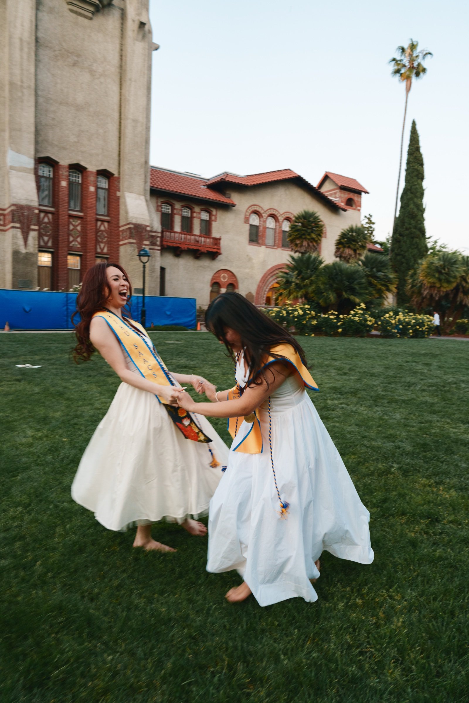 Two SJSU SAACS graduates in white dresses laughing and spinning barefoot on the grass in front of Torre Hall at sunset