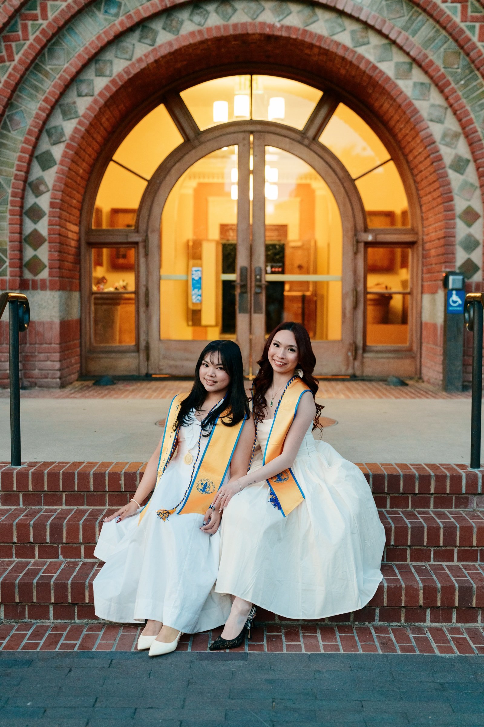 Two SJSU graduates in gold sashes sitting side by side on brick steps in front of the glowing arched Event Center doorway