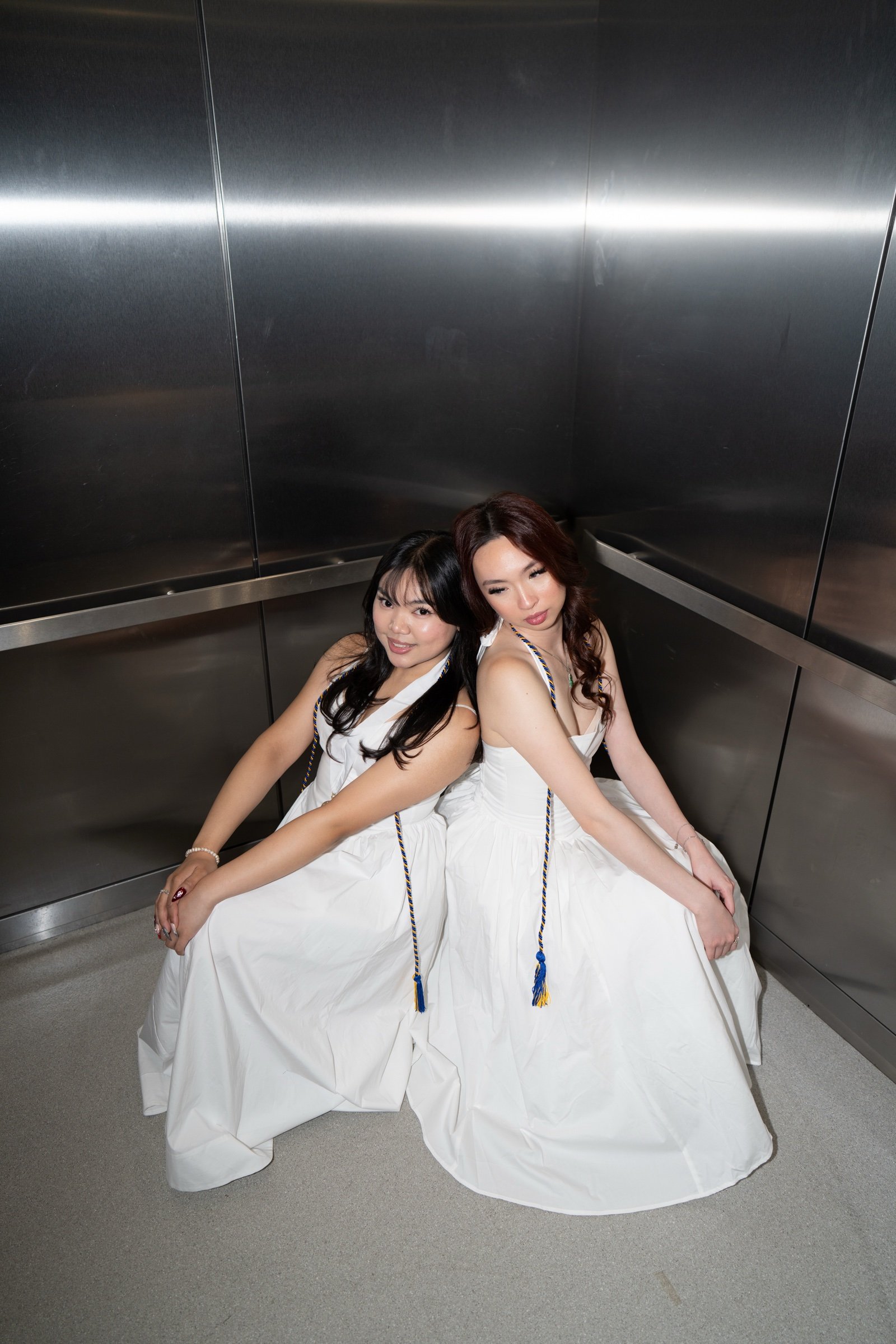 Two SJSU graduates in white dresses and honor cords sitting back-to-back on the floor of a stainless steel elevator