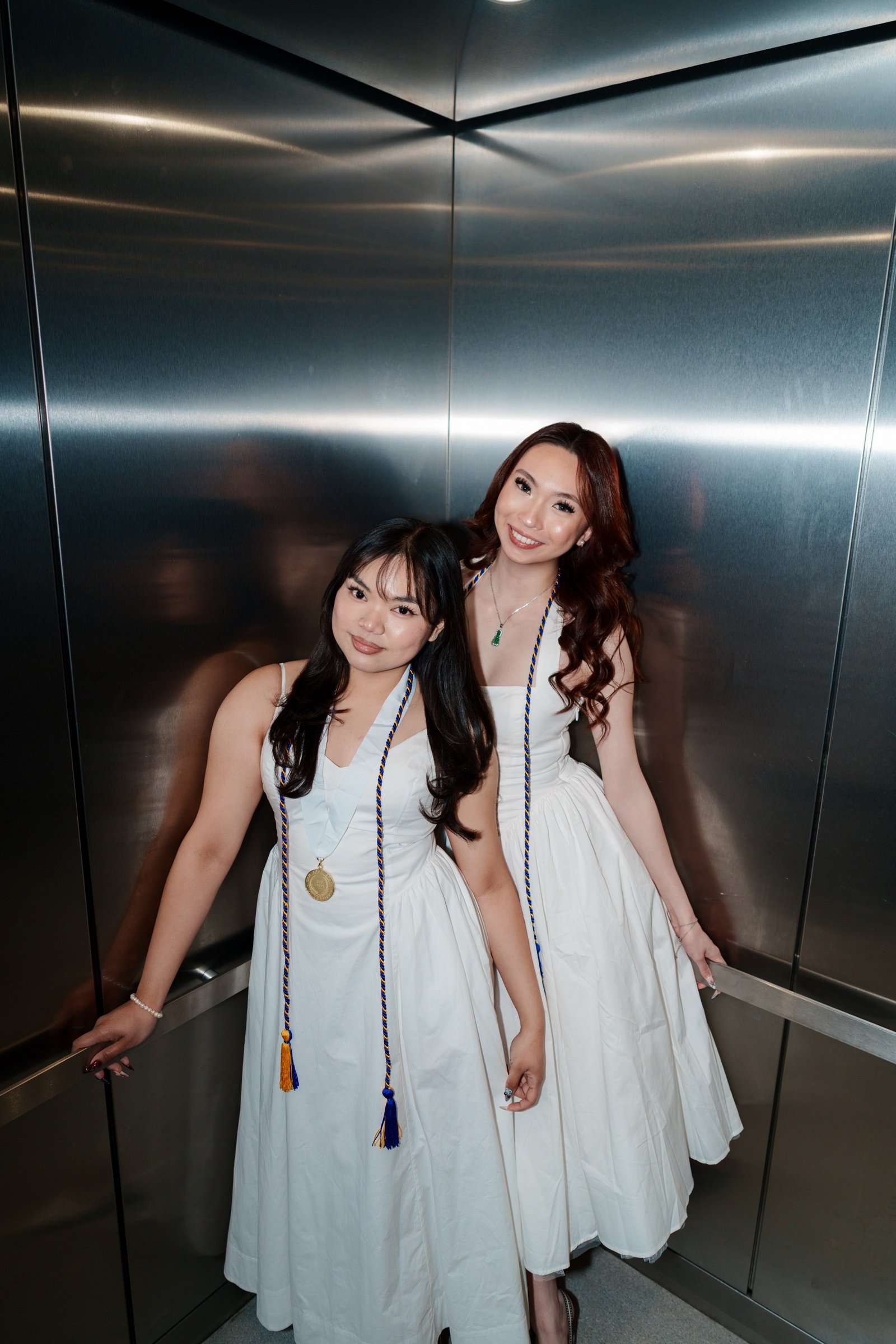 Two SJSU graduates in white dresses and blue-and-gold honor cords posing together inside a stainless steel elevator