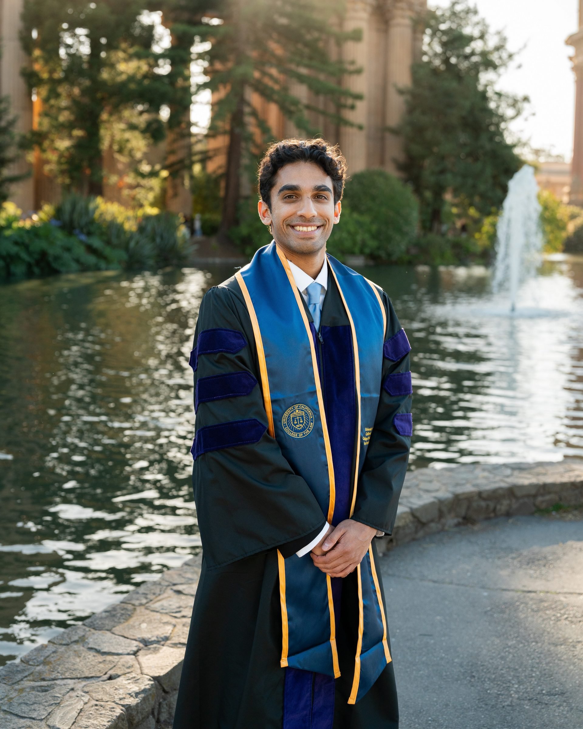 UC Law SF graduate stands smiling in doctoral regalia with blue and gold stole beside the lagoon at Palace of Fine Arts, a fountain and warm golden-hour light filling the background