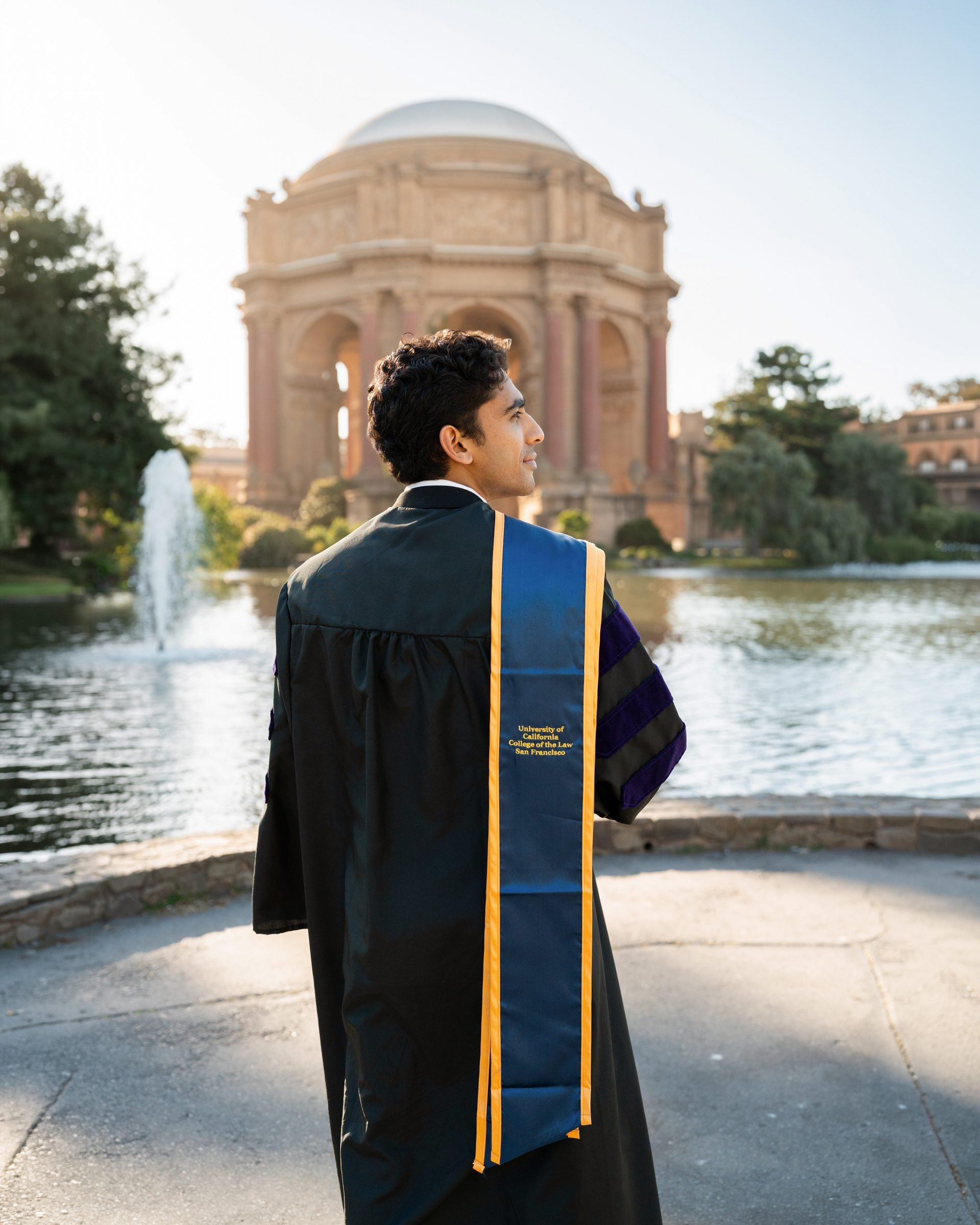 UC Law SF graduate in black doctoral regalia with blue and gold hood stands at the Palace of Fine Arts rotunda during golden hour, the lagoon fountain and warm sandstone dome glowing behind him