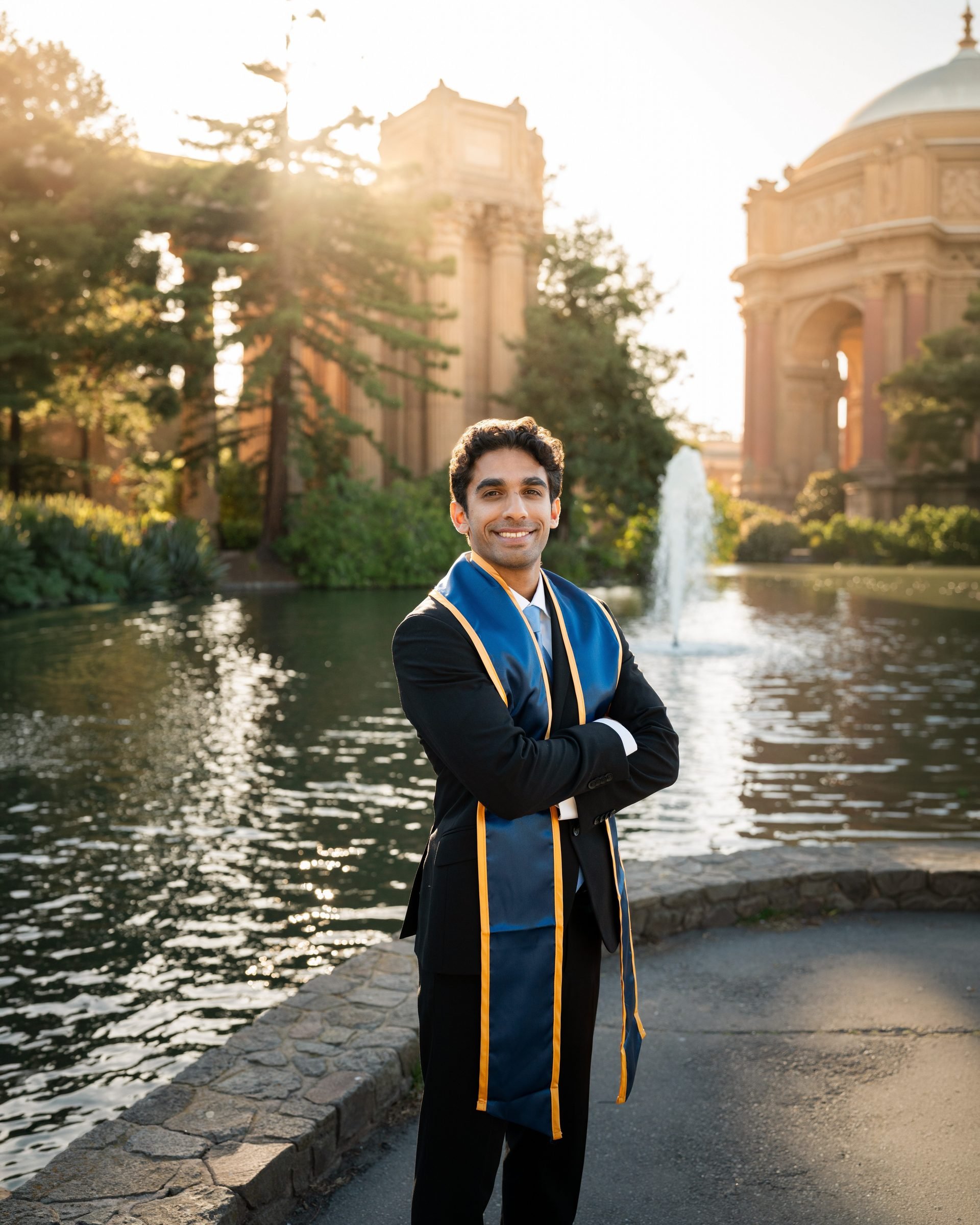 UC Law SF graduate in black suit and navy-and-gold stole stands arms crossed beside the Palace of Fine Arts lagoon, golden hour sunlight streaming through the colonnades behind him with the fountain visible in the background