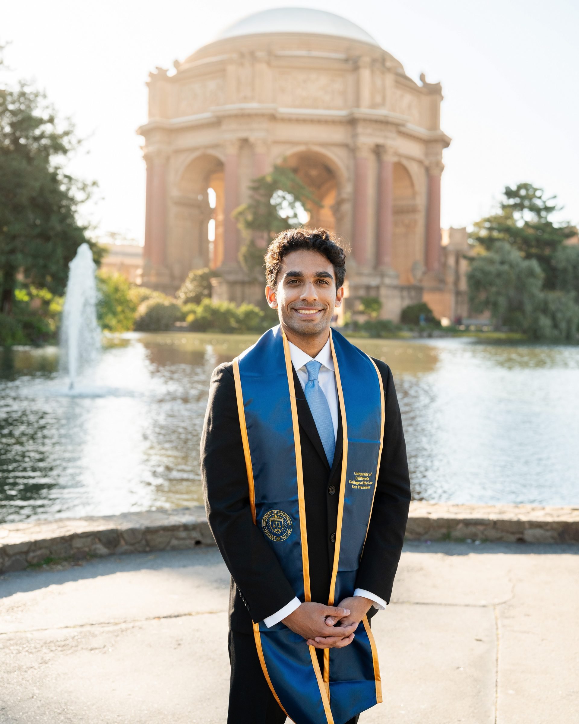 UC Law SF graduate smiling in a black suit and blue-and-gold stole, standing before the Palace of Fine Arts rotunda and lagoon fountain at golden hour in San Francisco
