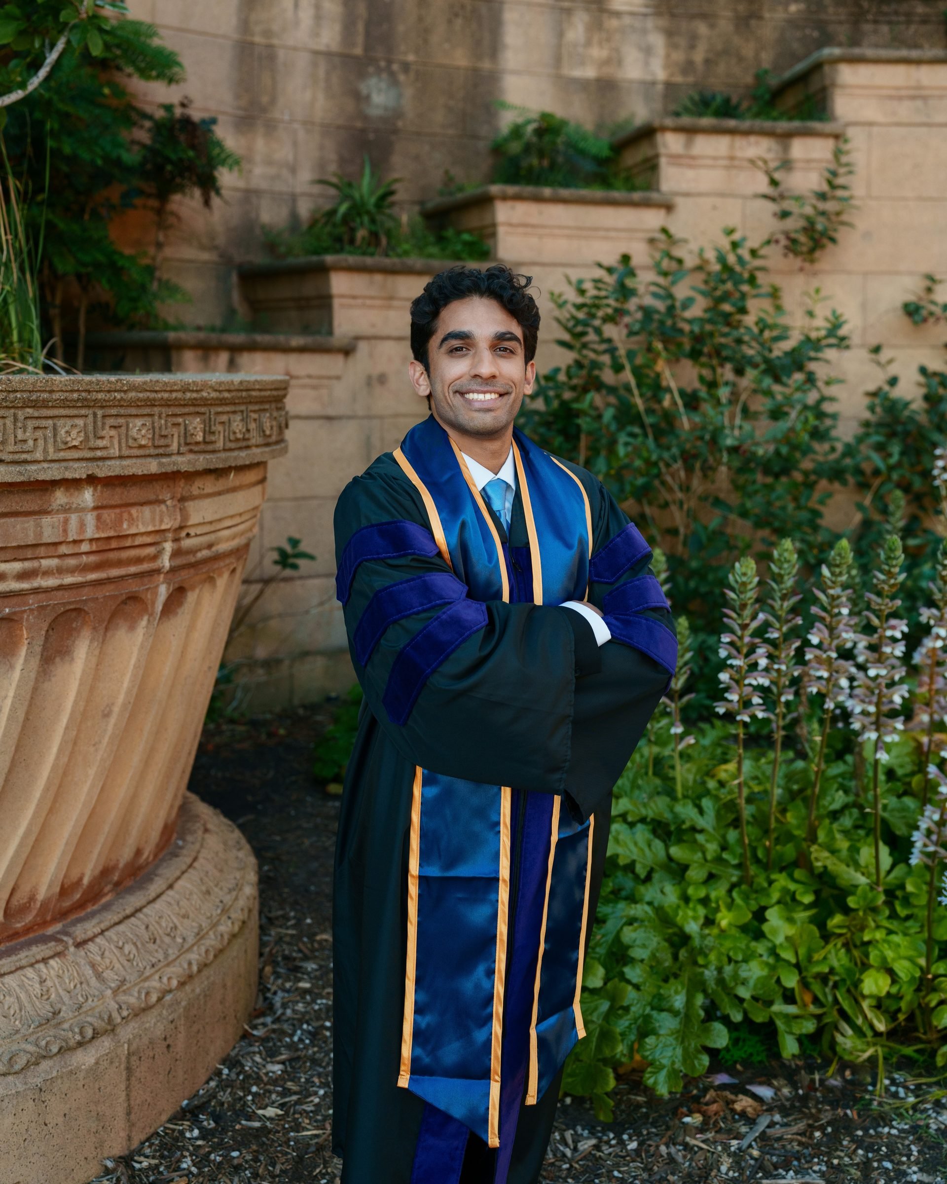 UC Law SF graduate stands arms crossed beside a large terracotta urn at the Palace of Fine Arts, smiling confidently in a dark doctoral gown with royal blue velvet and gold trim, surrounded by lush garden greenery in warm golden-hour light.