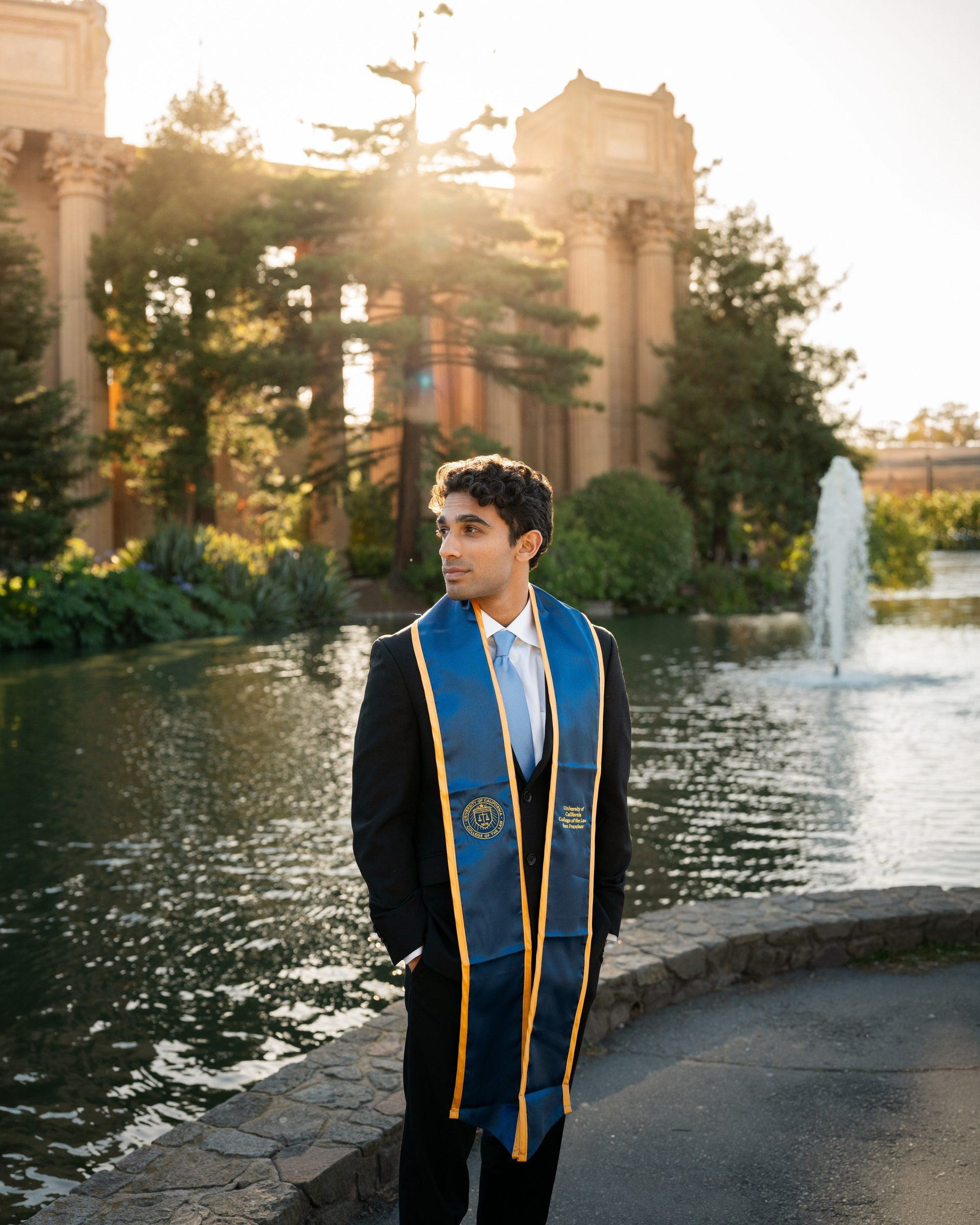 UC Law SF graduate in a black suit and blue-and-gold stole stands beside the lagoon at the Palace of Fine Arts, golden-hour sun flaring through the rotunda columns behind him