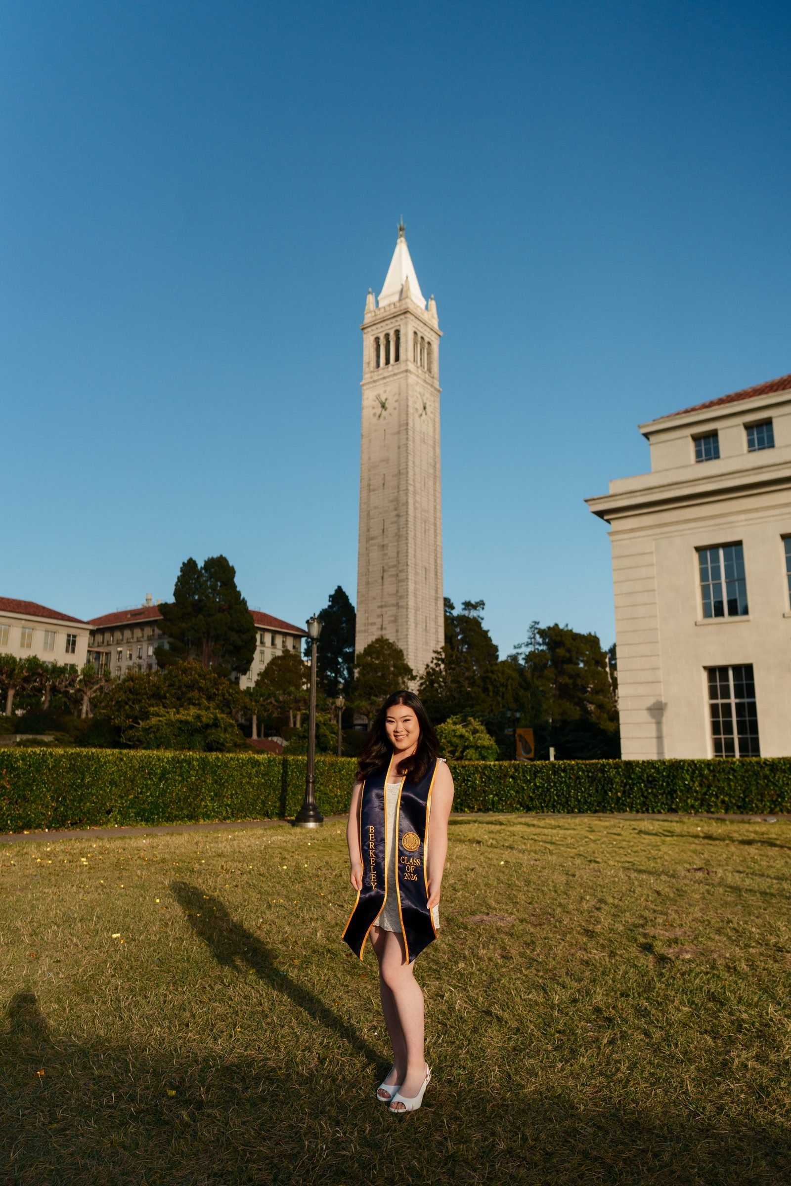UC Berkeley grad Kim stands on Memorial Glade smiling with Sather Tower rising behind her, wearing a white dress and Berkeley Class of 2026 sash, bathed in warm low-angle golden-hour sun with long shadows across the lawn.