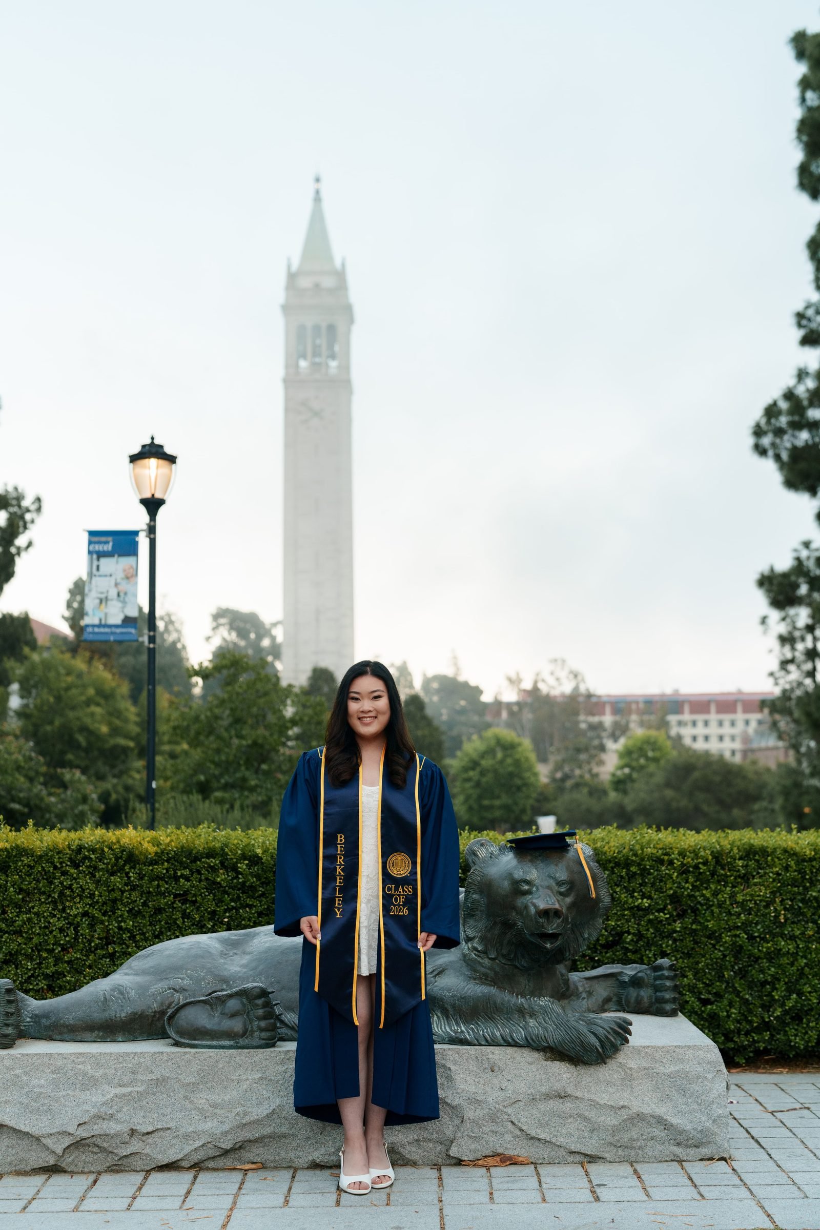 Kim stands in front of the Cal Bear statue in navy graduation gown and Berkeley stole, both hands holding the gown open, with Sather Tower and green campus trees framing the scene behind her.