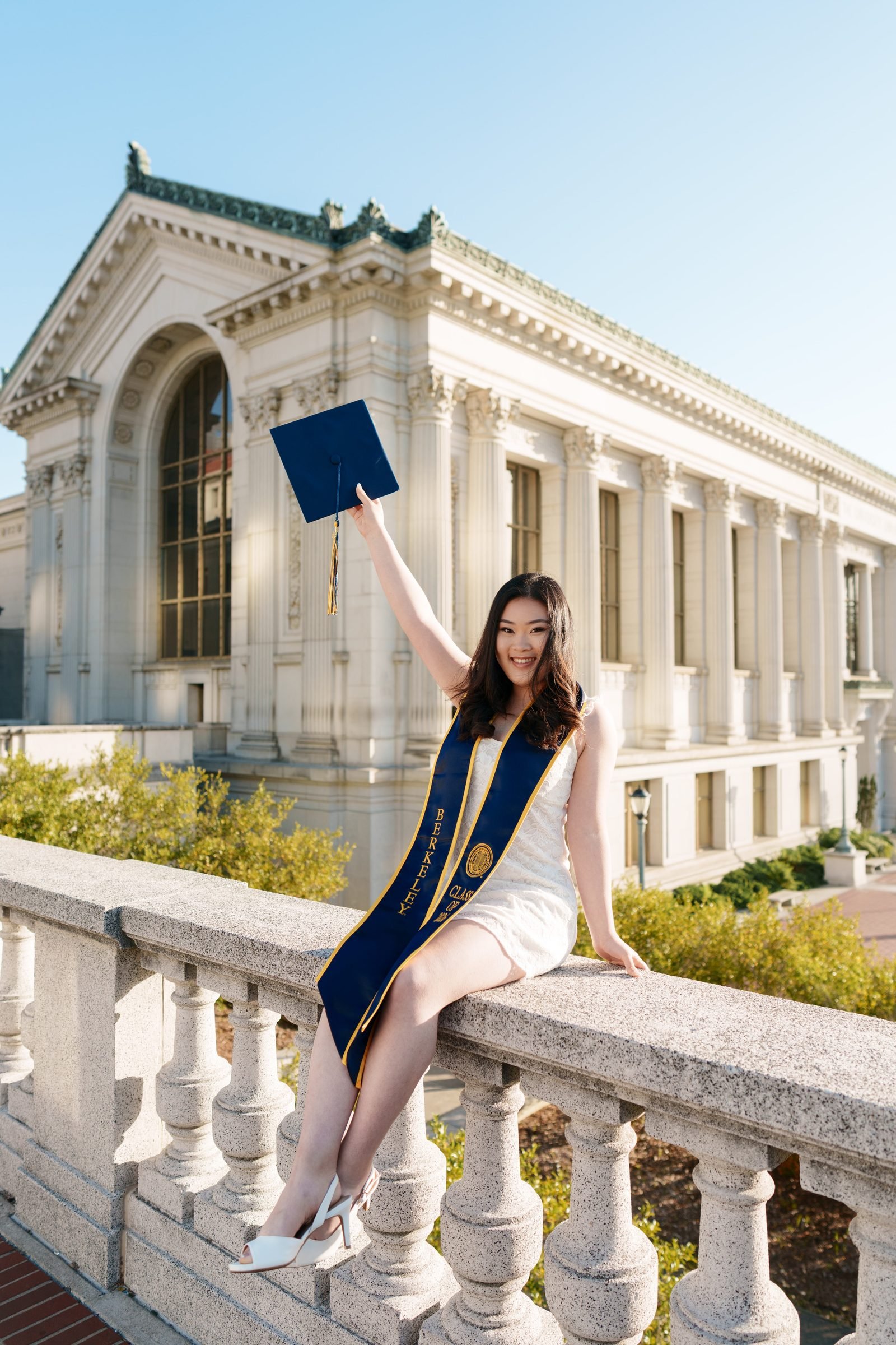 UC Berkeley grad Kim sits on a stone balustrade in front of Doe Library, arm raised holding her navy mortarboard, wearing a white dress and Berkeley Class of 2026 sash in warm golden-hour light.