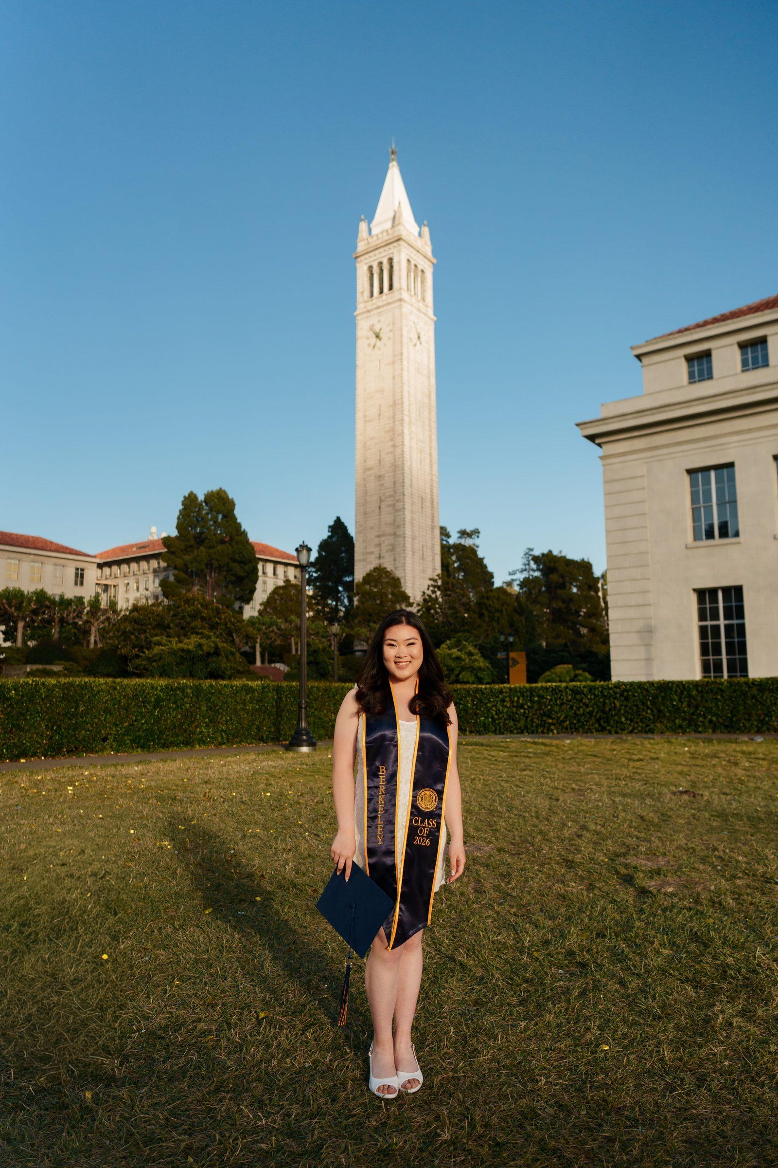 UC Berkeley grad Kim stands on the grass of Memorial Glade holding her navy cap, with Sather Tower rising into a clear blue sky behind her, wearing a white dress and Berkeley Class of 2026 sash in golden-hour light.