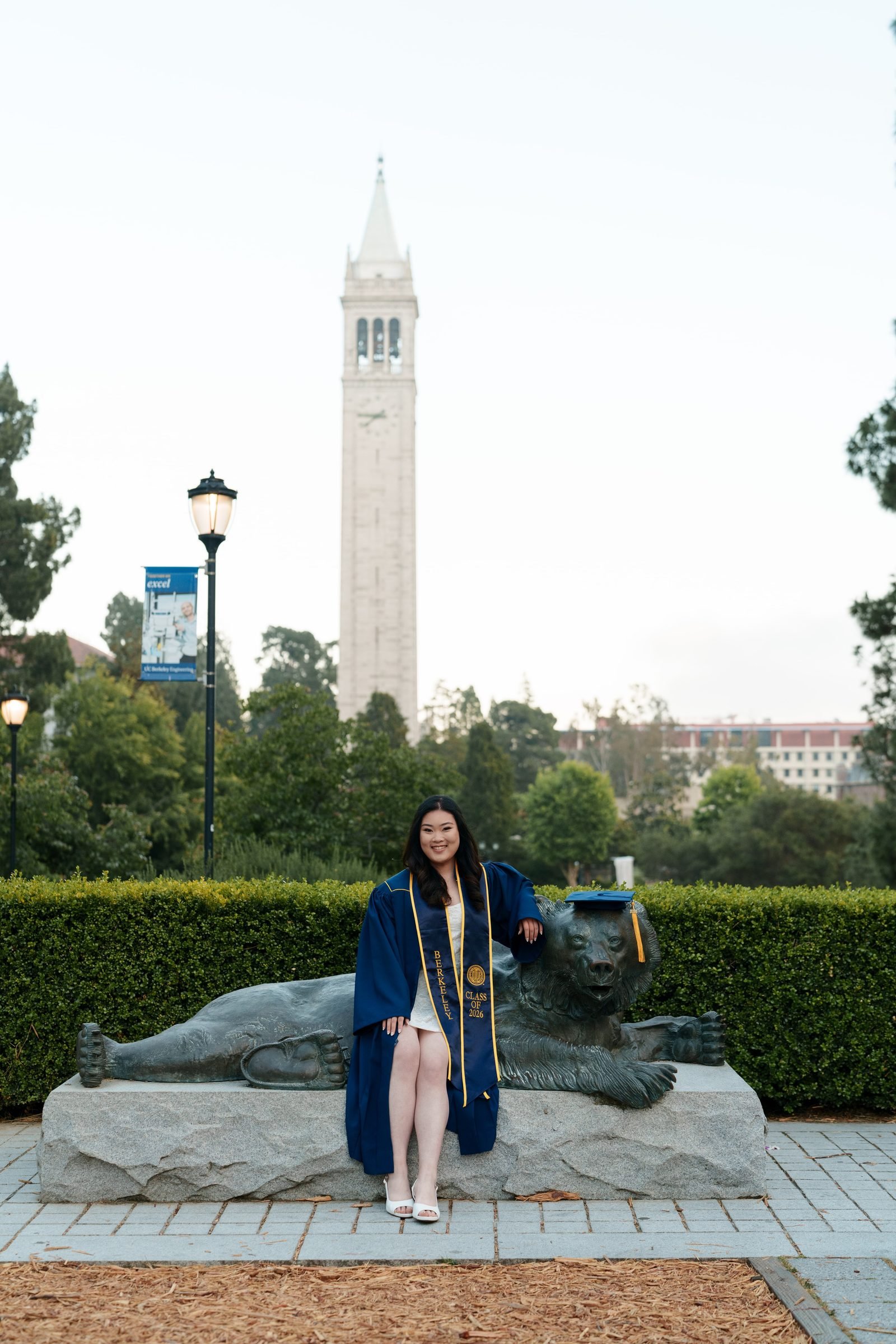 Berkeley grad Kim sits on the Cal Bear statue in navy cap and gown with gold Berkeley stole, Sather Tower rising in the soft evening sky behind her, white dress and heels peeking out beneath her robe.