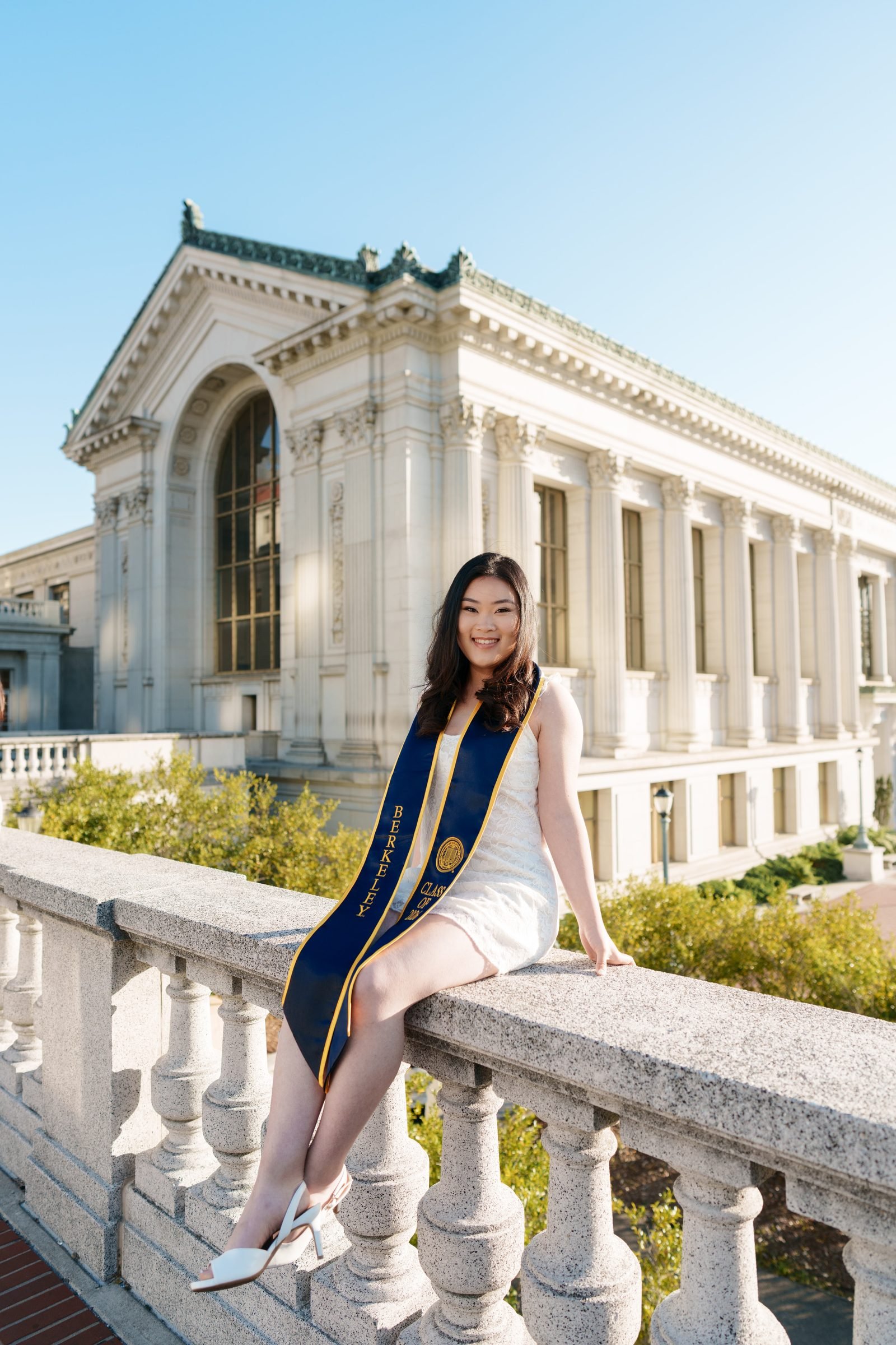 Kim sits on a stone balustrade in a white dress and navy Berkeley stole, Doe Library's Beaux-Arts facade filling the background under a clear blue sky, morning light casting soft shadows across the scene