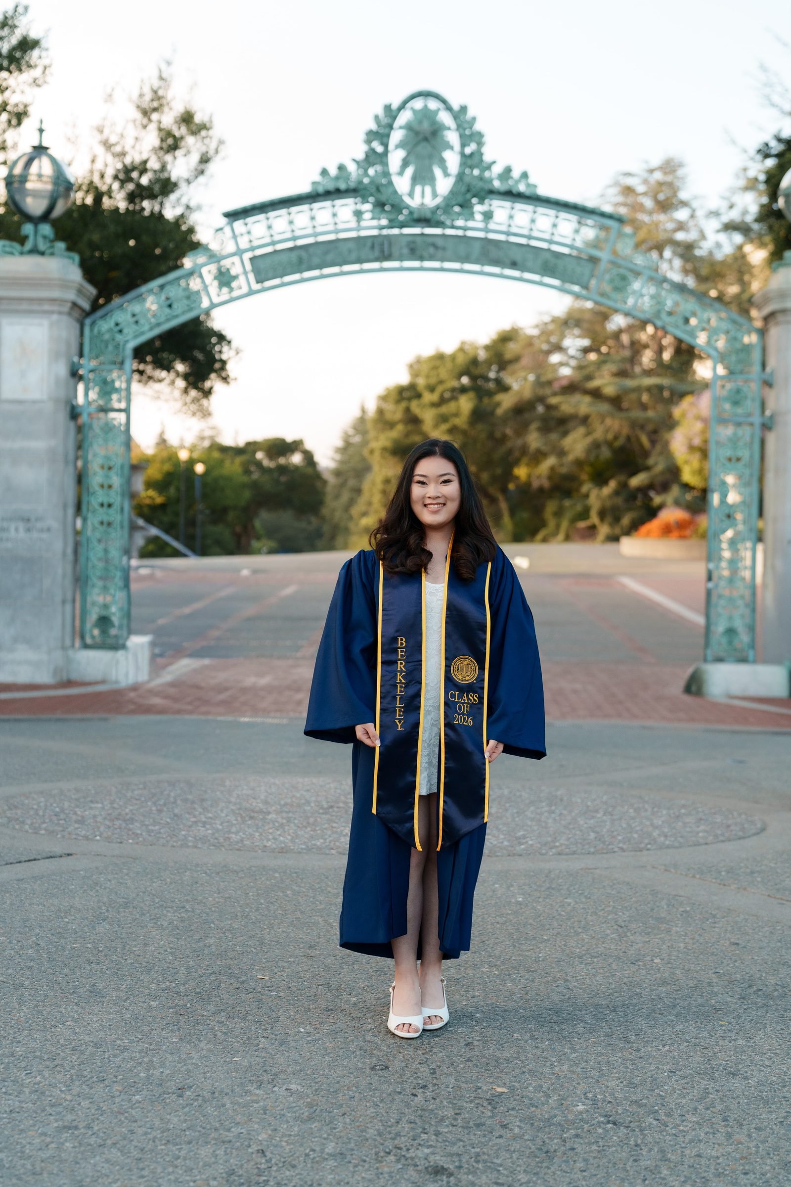 Kim stands in front of Sather Gate in a full navy graduation gown and Berkeley sash, smiling at camera during soft golden-hour light with the ornate teal metalwork arch filling the background.