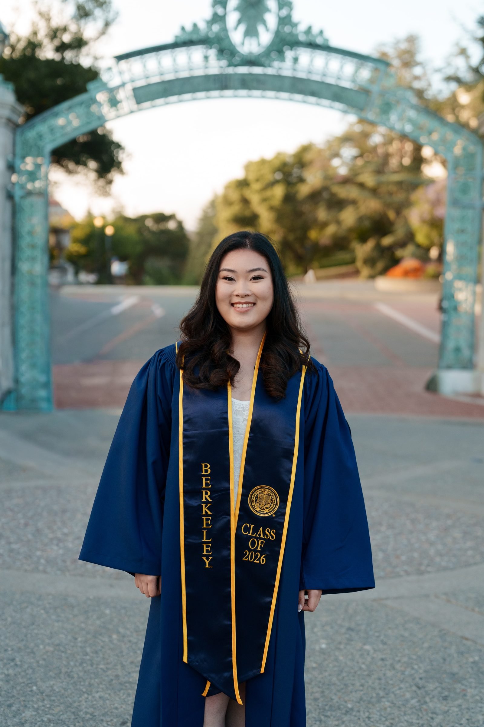 Close portrait of UC Berkeley grad Kim in navy gown and gold Berkeley stole, smiling brightly in front of Sather Gate with soft golden-hour bokeh in the background