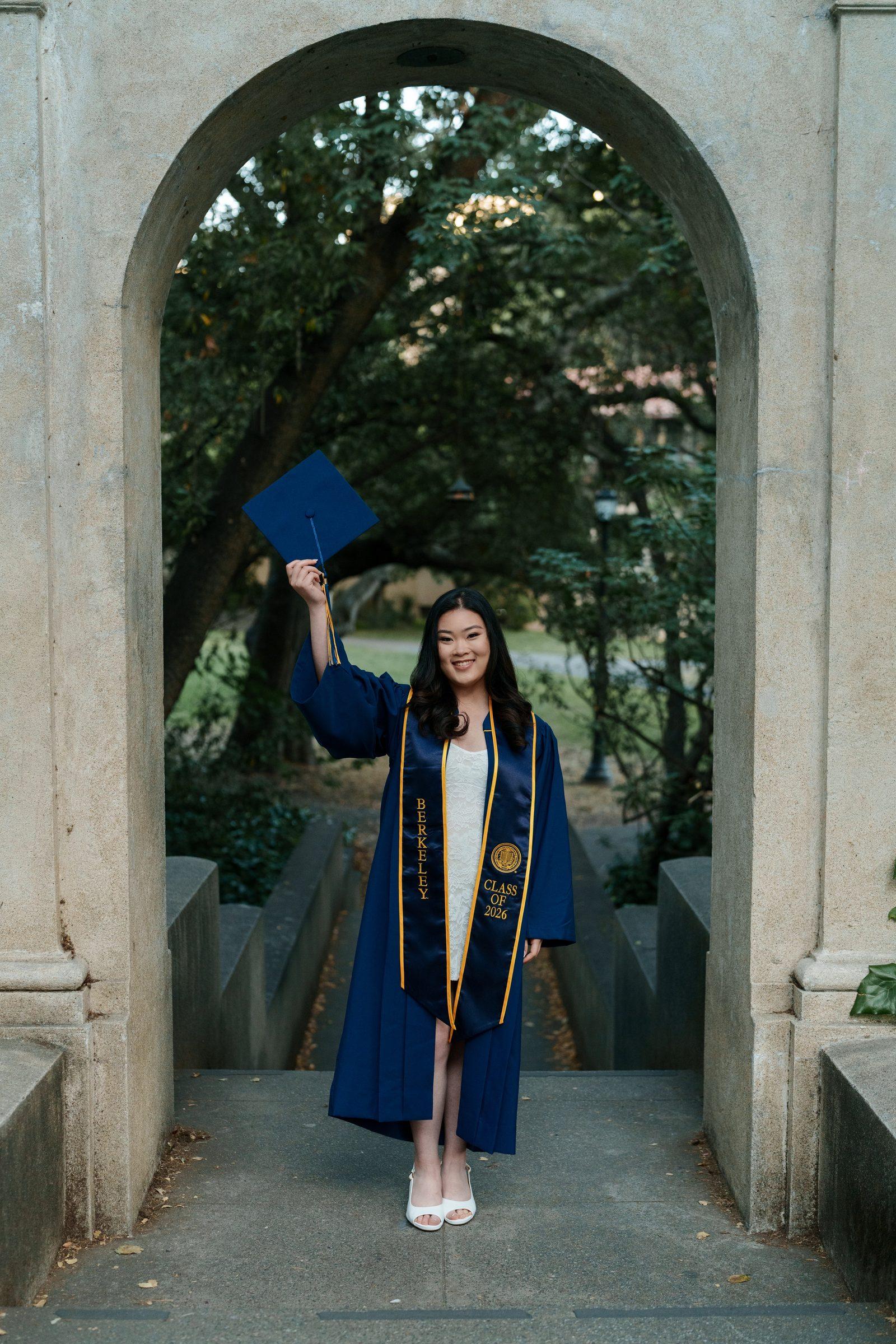 Kim raises her navy graduation cap under a stone archway on the UC Berkeley campus, wearing her graduation gown and Berkeley stoles, smiling warmly in soft ambient evening light.