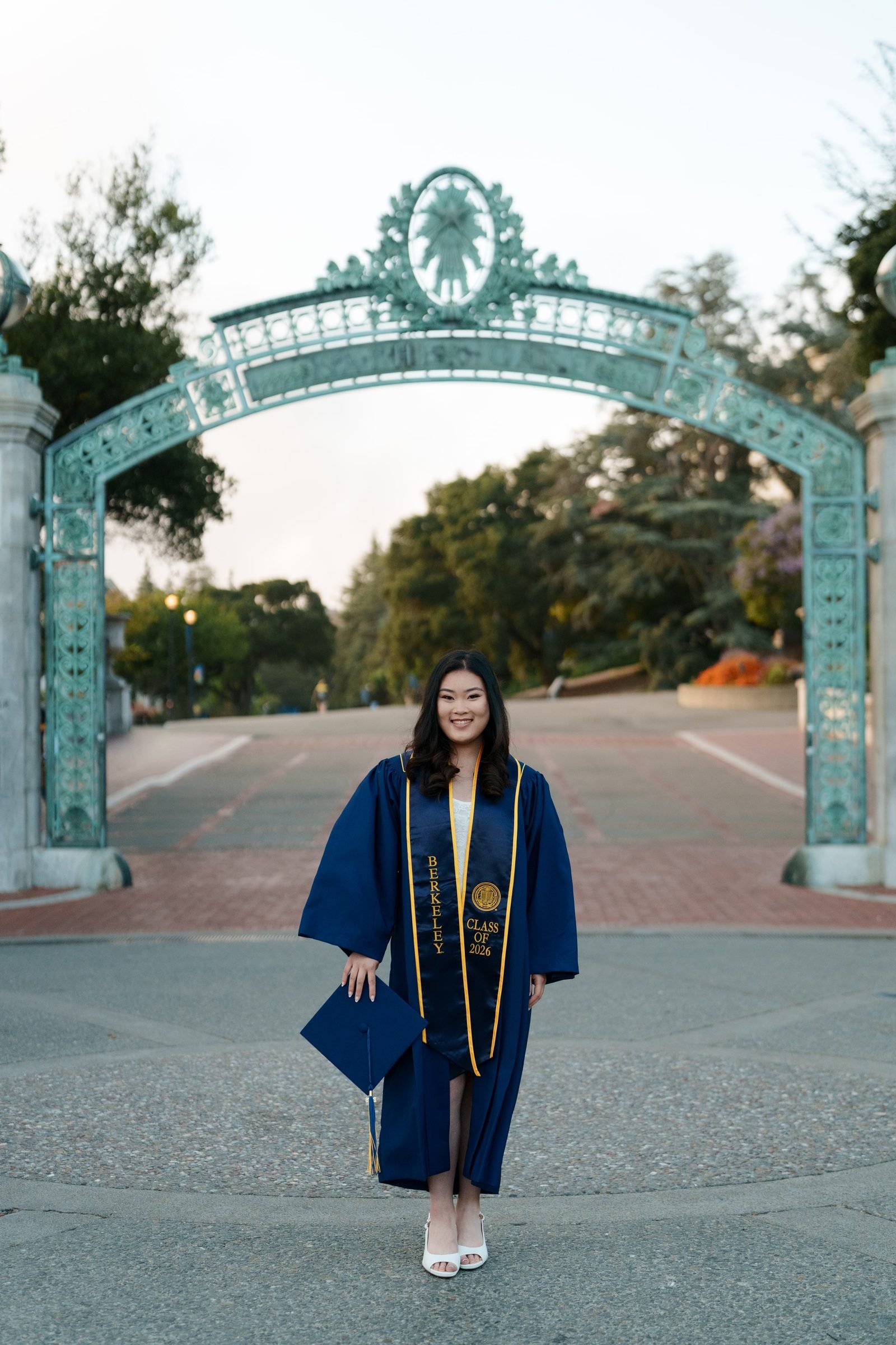 UC Berkeley grad Kim walks toward camera in navy cap and gown with gold stole, centered under Sather Gate, smiling confidently in soft early-evening light with campus path behind her