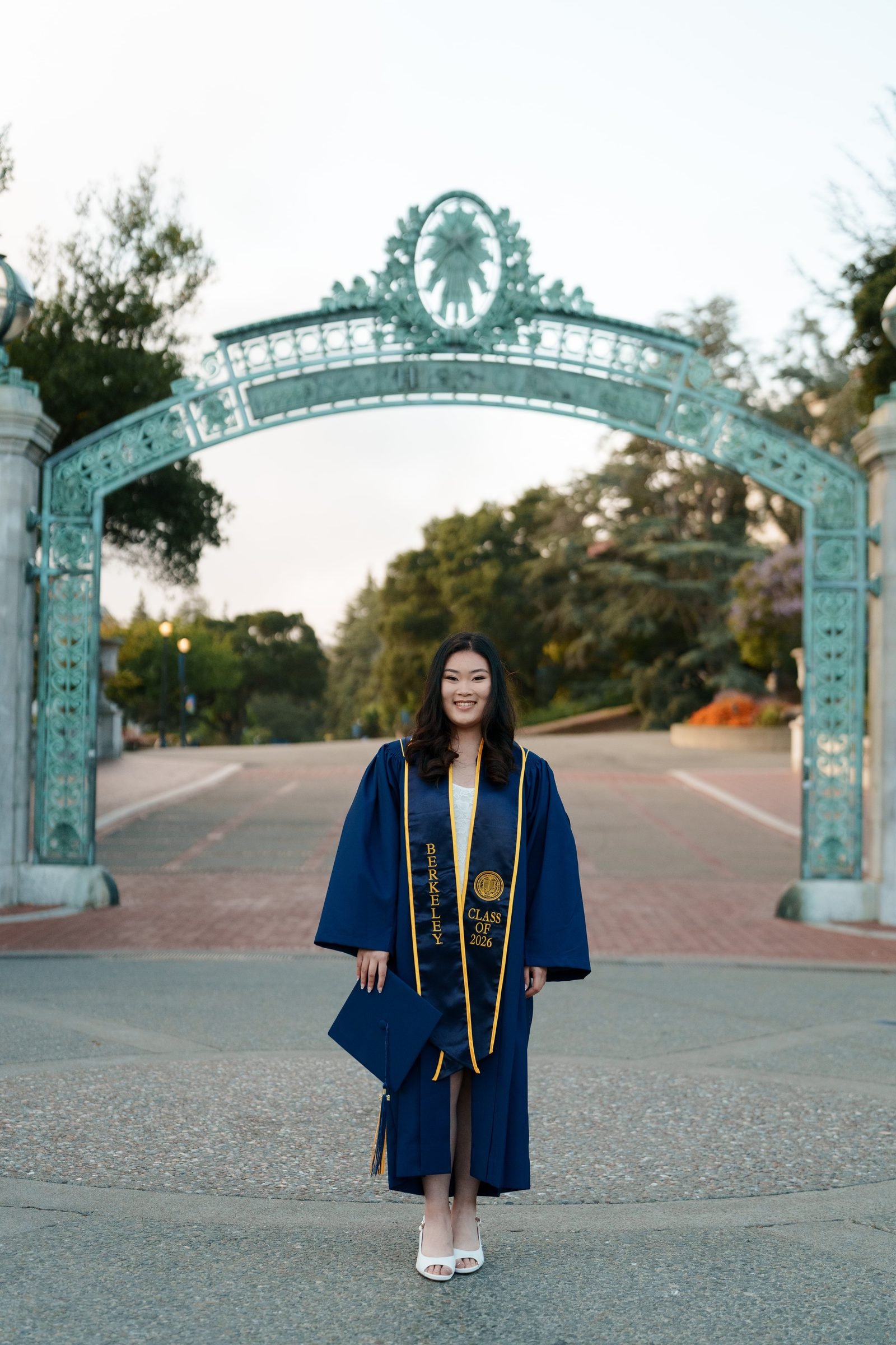 UC Berkeley grad Kim stands centered beneath Sather Gate in navy cap and gown with gold Class of 2026 stole, holding her cap at her side, smiling warmly in soft evening light