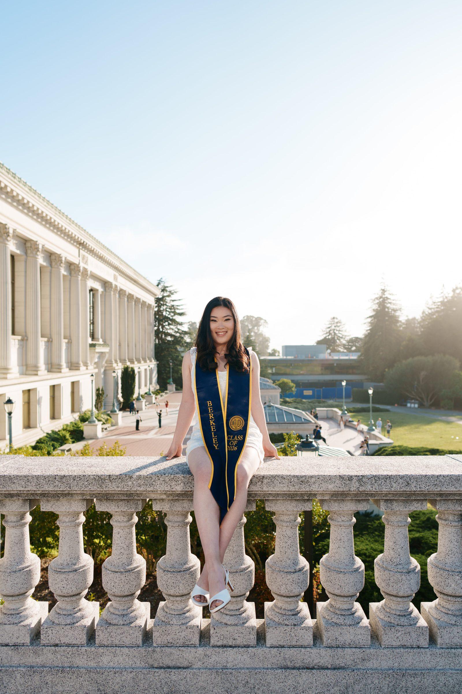 Kim sits relaxed on a stone balcony railing in a white dress and Berkeley Class of 2026 stole, golden backlighting creating a warm haze, with Doe Library visible in the background.