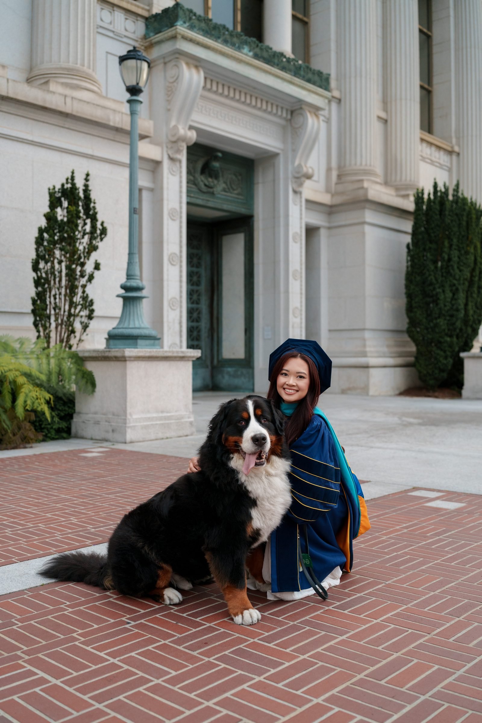 Graduate in blue doctoral regalia crouches beside a happy Bernese Mountain Dog on brick pavers in front of a grand stone building with tall columns and ornate detailing at Doe Library.