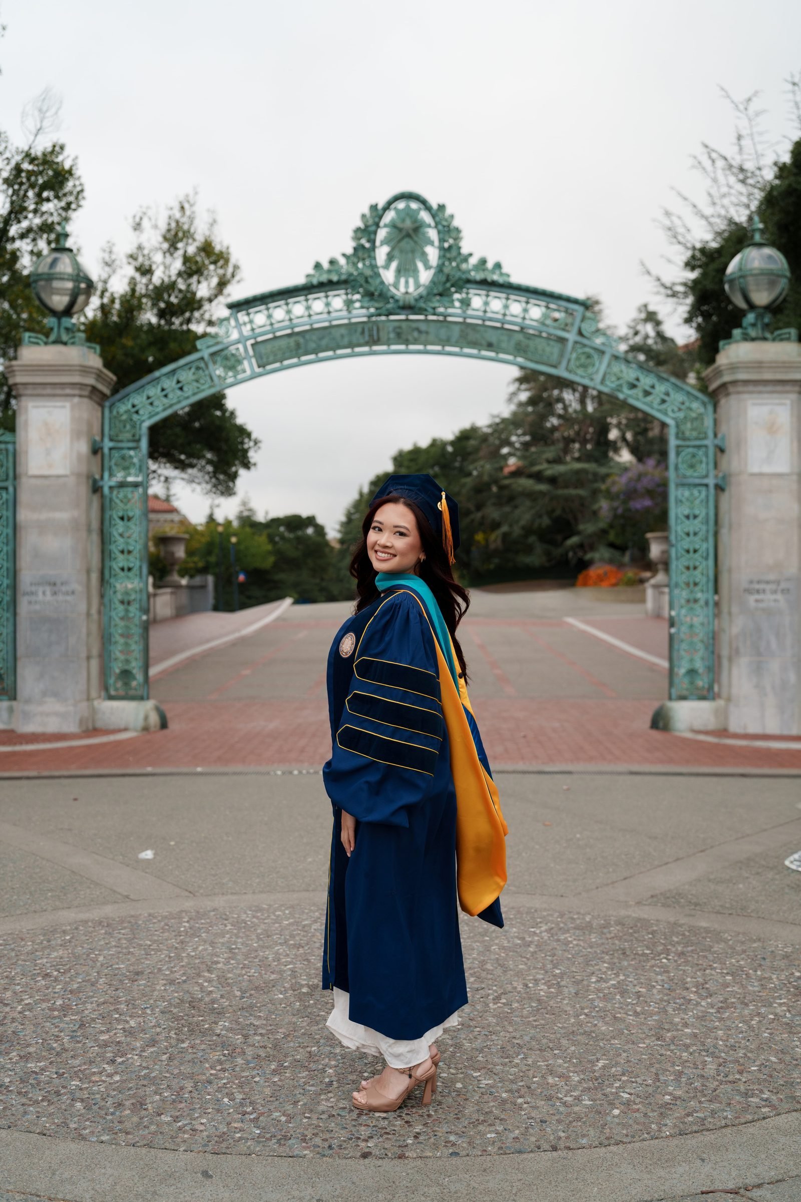 Trang stands in front of Sather Gate in a navy doctoral gown with gold trim and teal hood, glancing back over her shoulder with a warm smile on a softly overcast morning.