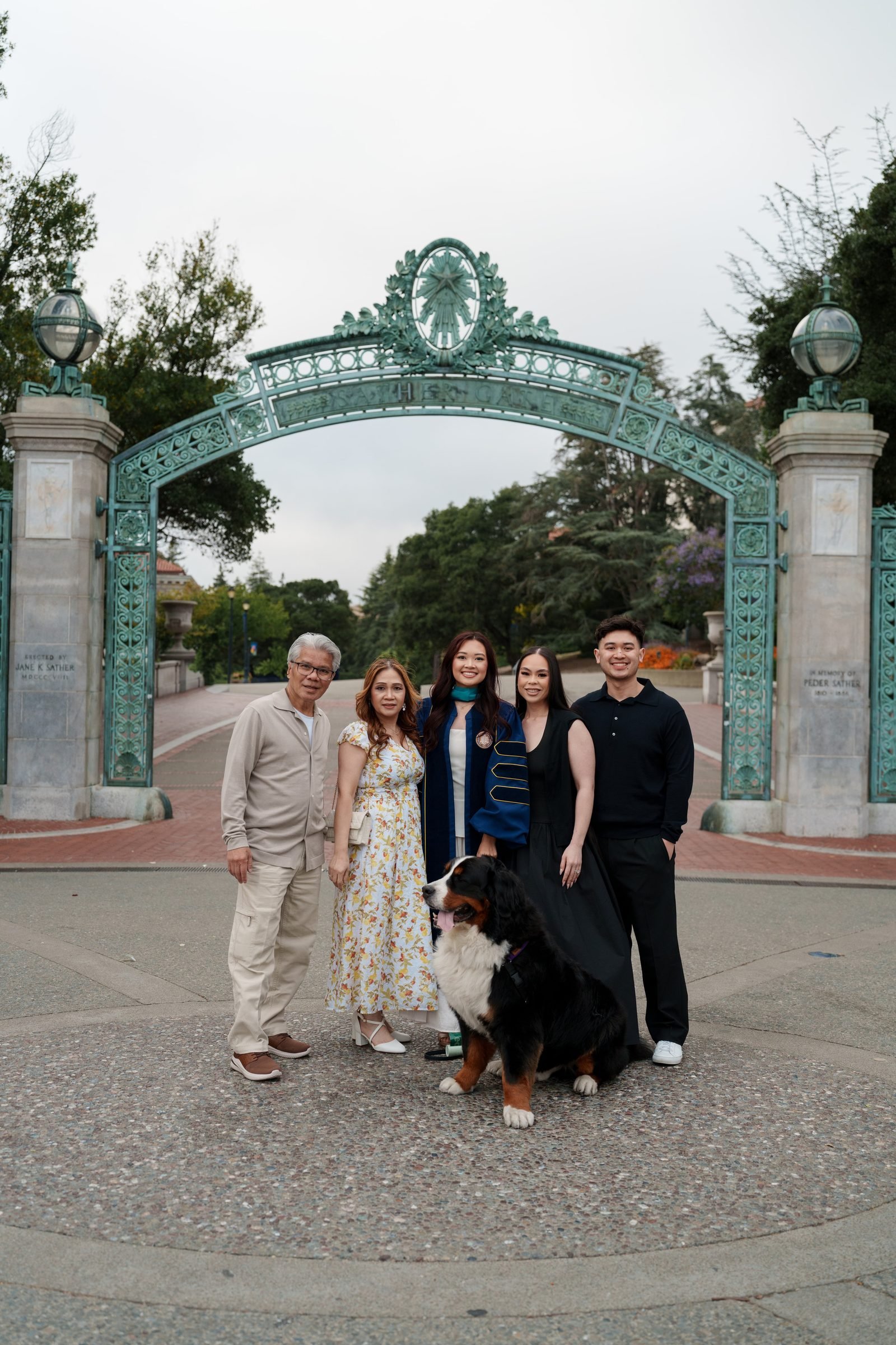 Family of five plus a Bernese Mountain Dog posed in front of Sather Gate at UC Berkeley under overcast light; graduate wears navy regalia with a teal hood while family members wear neutral and floral outfits
