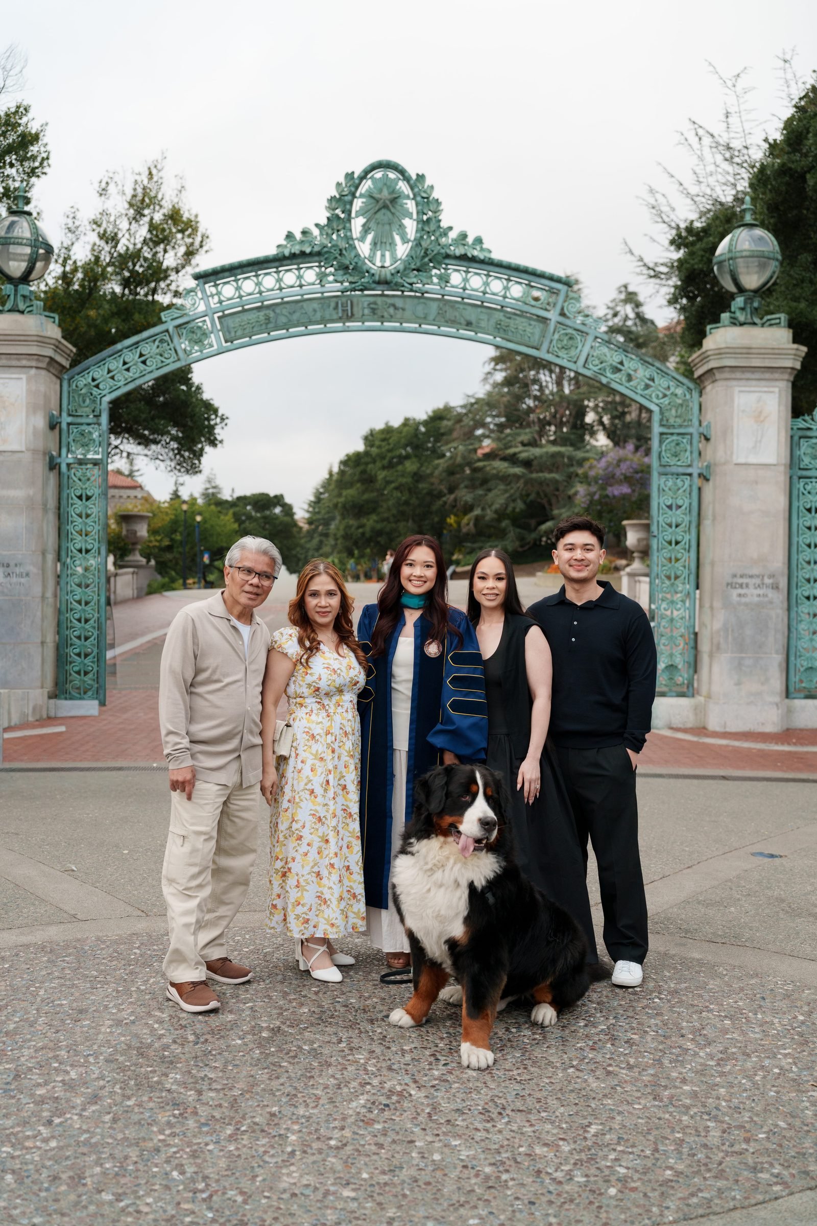 A graduate in a navy blue doctoral gown stands with four family members and a Bernese Mountain Dog under the iconic teal Sather Gate at UC Berkeley on a soft overcast day.