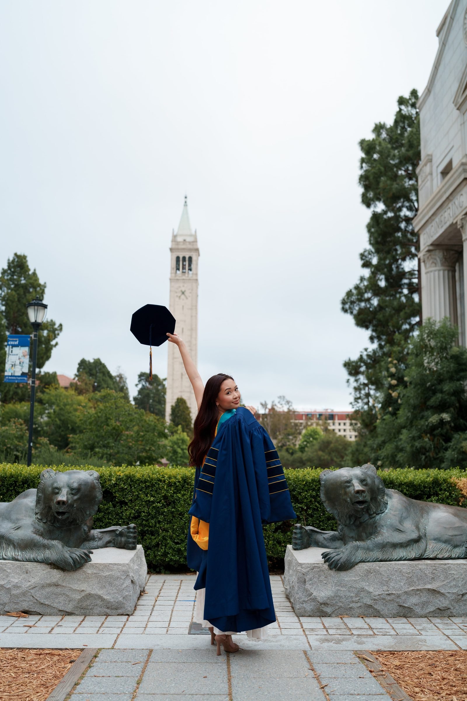 Graduate in navy doctoral regalia raises her cap skyward between two bronze bear statues, glancing back over her shoulder with a smile, Sather Tower framing the overcast background at UC Berkeley.